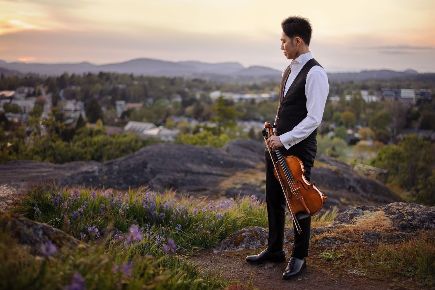 A man wearing an elegant vest watches the sunset at the edge of a mountain, holding a viola.