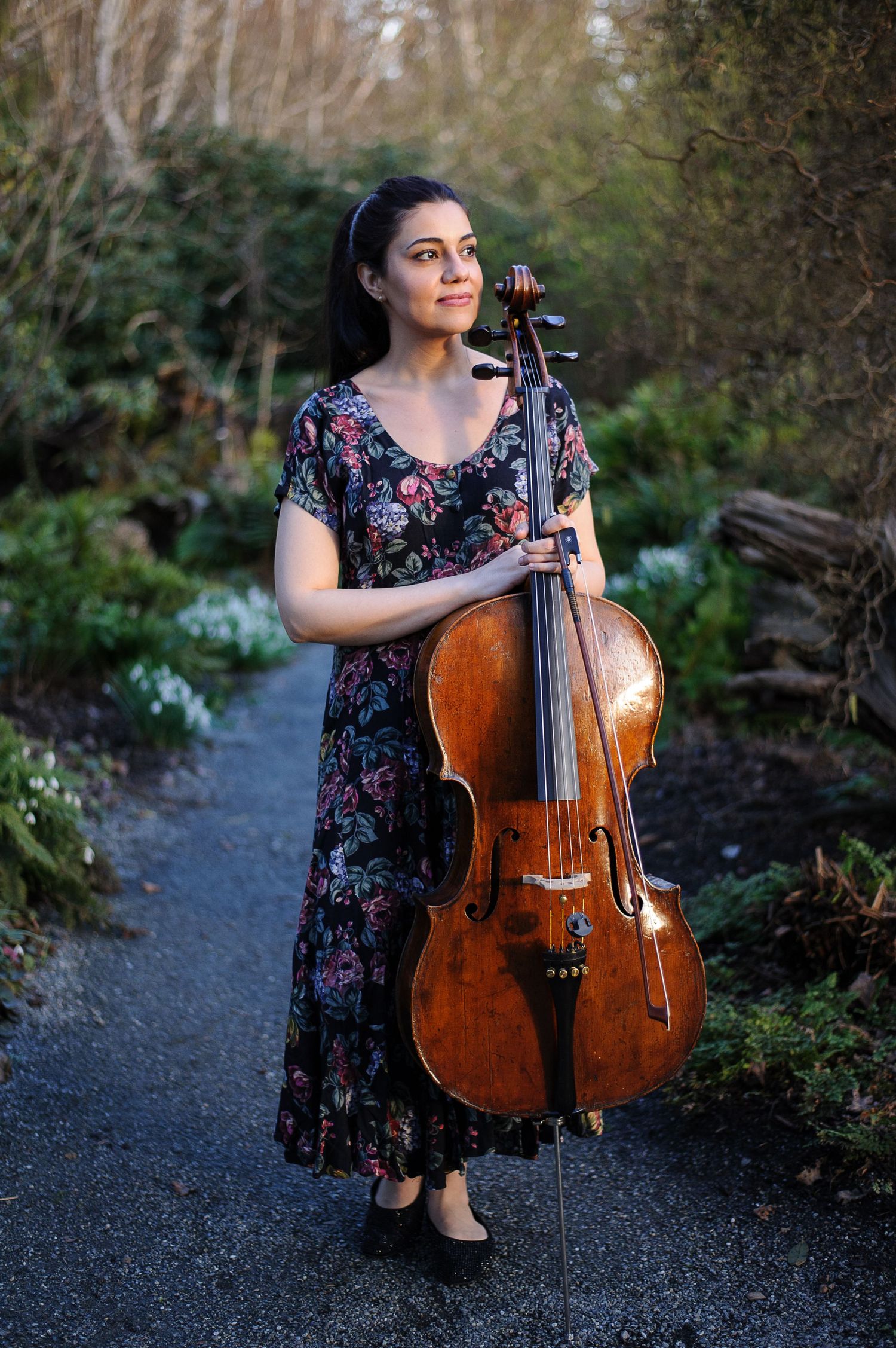 A woman in a flowery dress holds a cello and walks down a garden path.