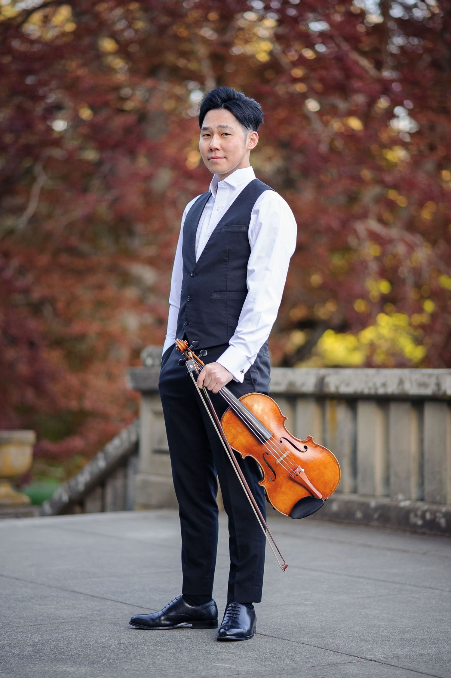 A man in an elegant vest strikes a pose and stands holding a viola on a landing by some red trees.
