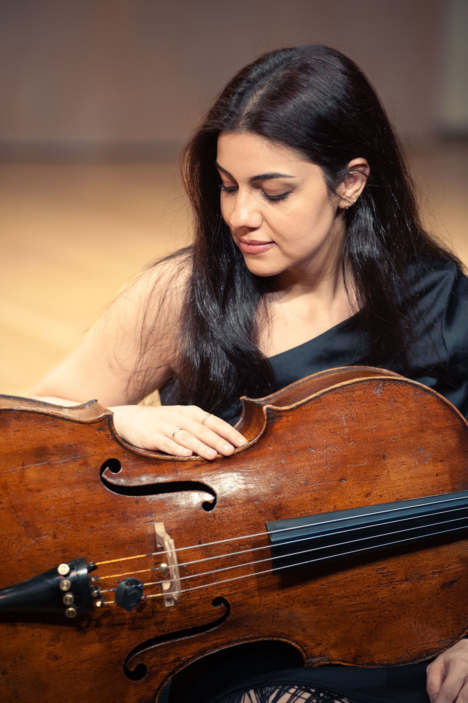 A woman in a black dress sits at the edge of a stage and lets her long black hair fall on the side of a cello.