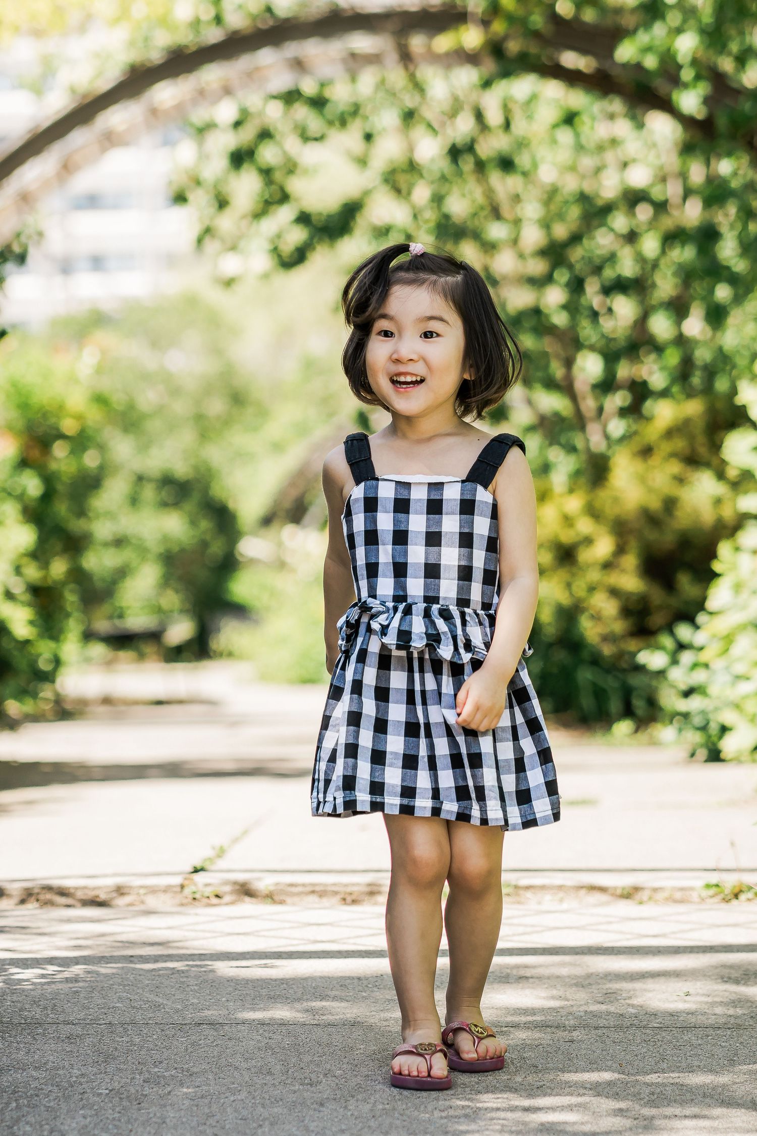 A young girl in a checkered sundress and pink flip flops walks through a leafy garden.