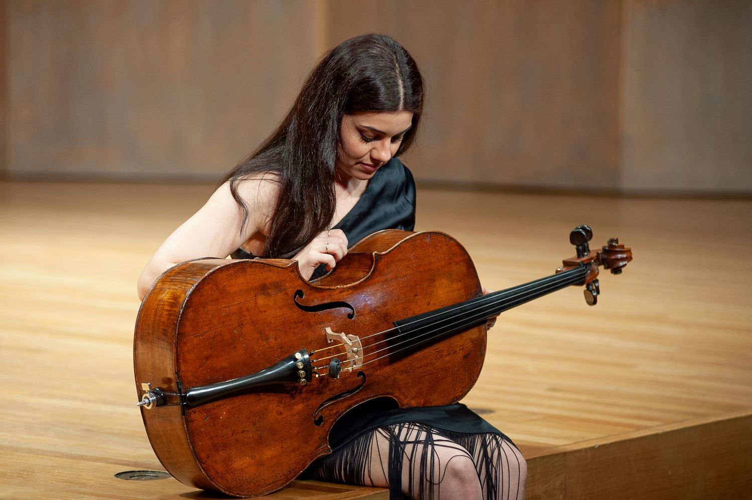 A woman in a black dress with long black hair sits at the edge of a stage holding her cello sideways.