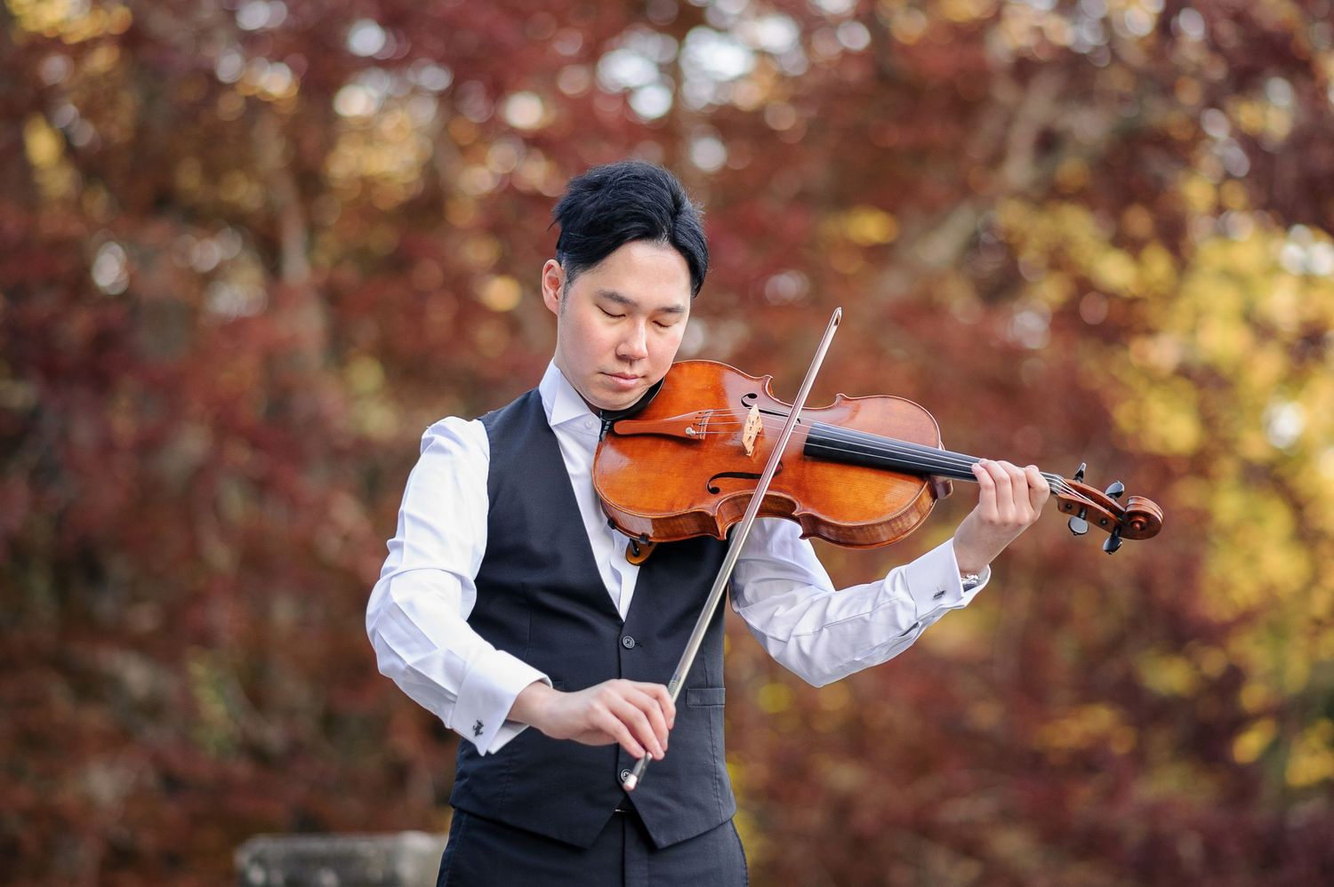 A man in an elegant vest plays viola in front of red and yellow leaves.