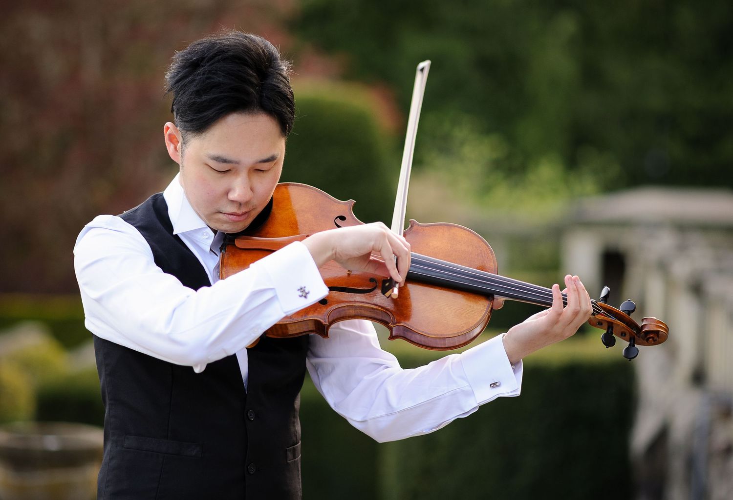 A man in an elegant vest plays viola in a garden.