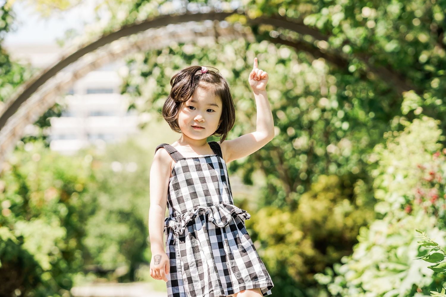 A young girl wearing a checkered sundress in a leafy garden, framed by an arch, points to the sky.