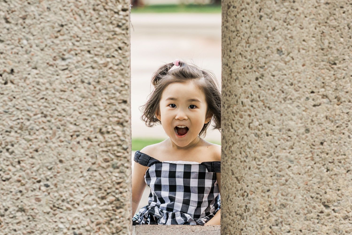 A young girl in a checkered sundress laughs and looks through a concrete opening.