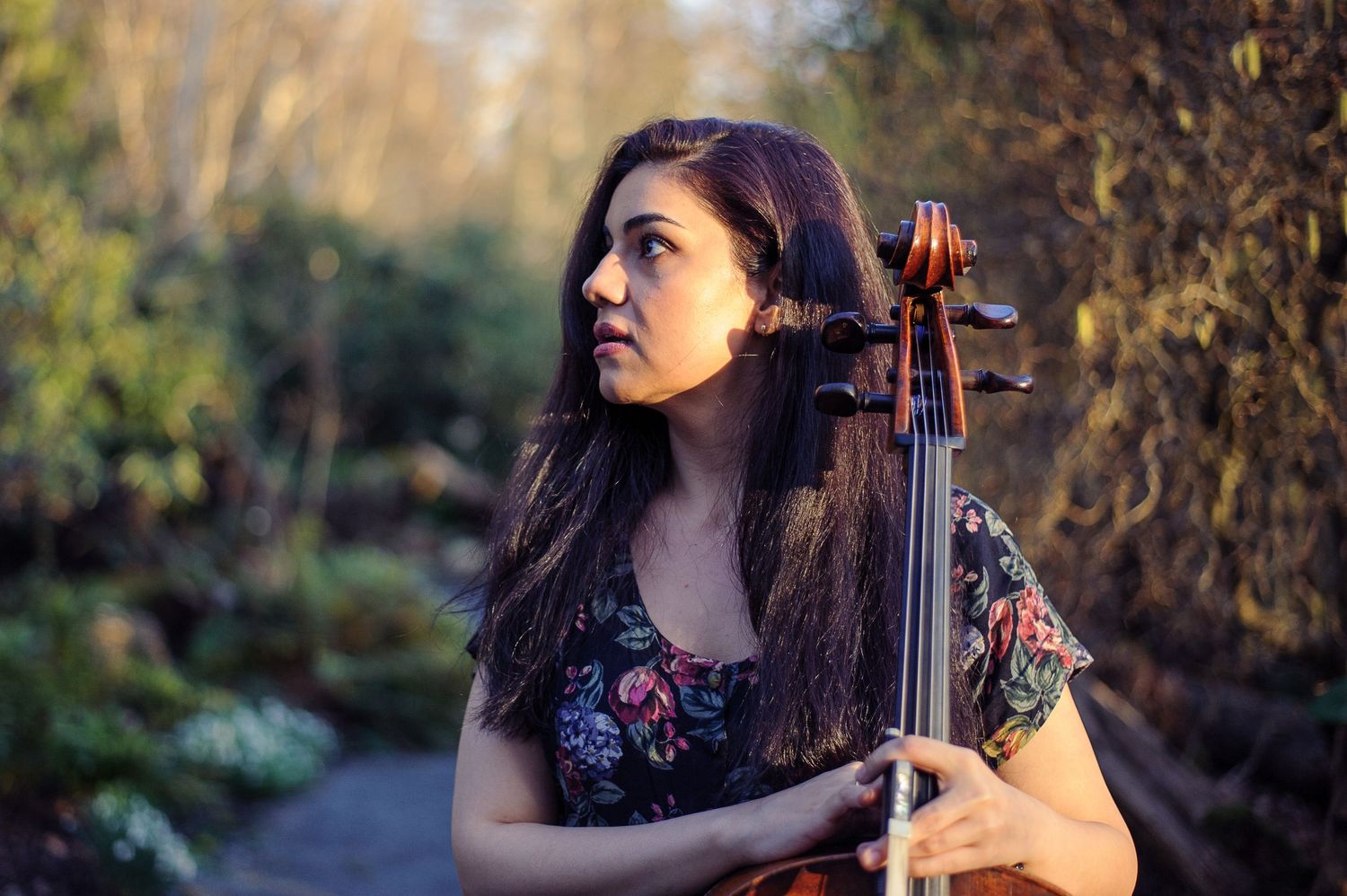A portrait of a woman holding a cello in a garden as she steps out of the shade.