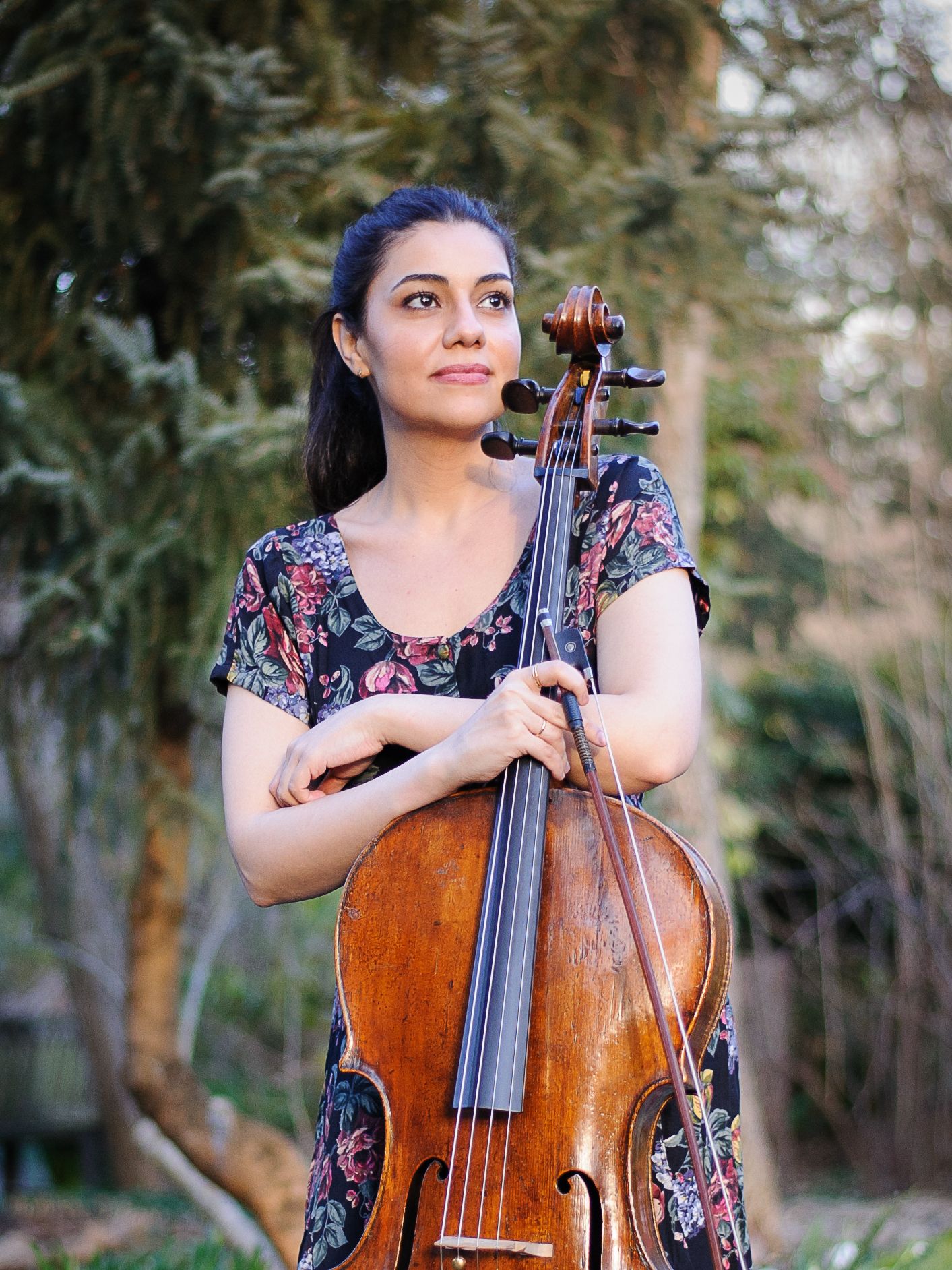 A woman in a flowery dress stands in front of pine trees and hugs her cello.