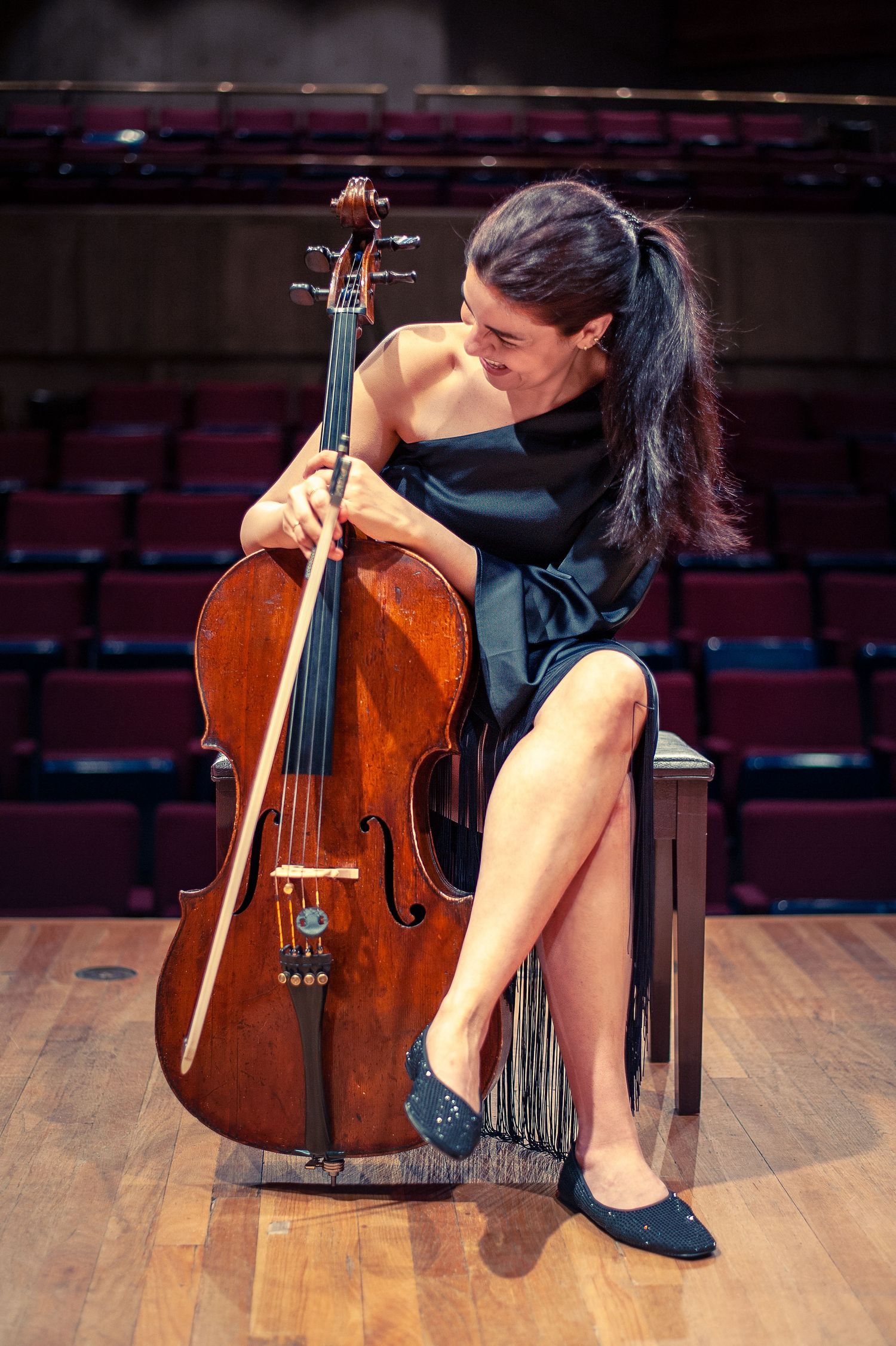 A woman sitting in a black dress on stage laughs and looks at her cello.