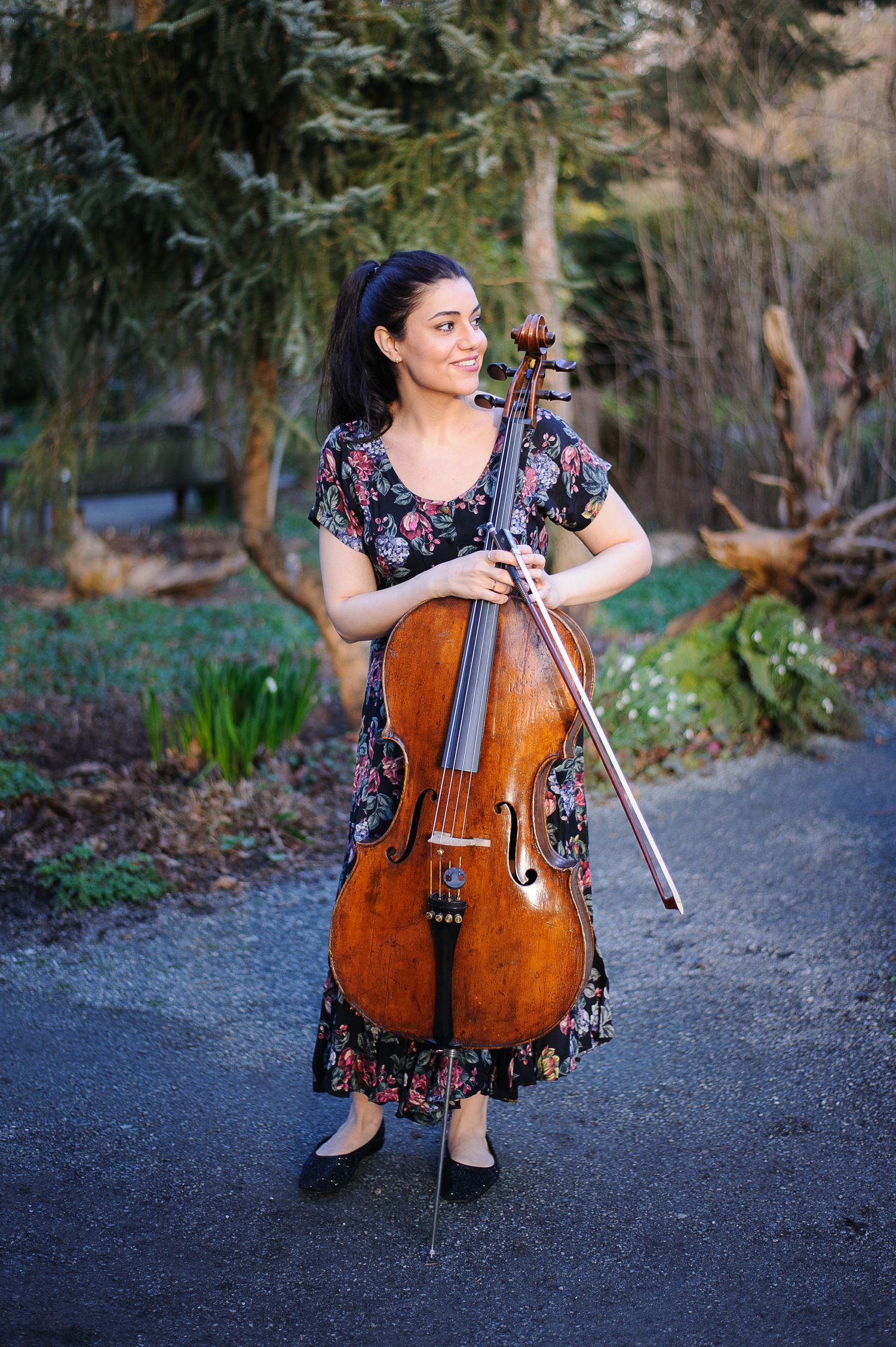 A woman in a flowery dress holds her cello and smiles on a garden path.