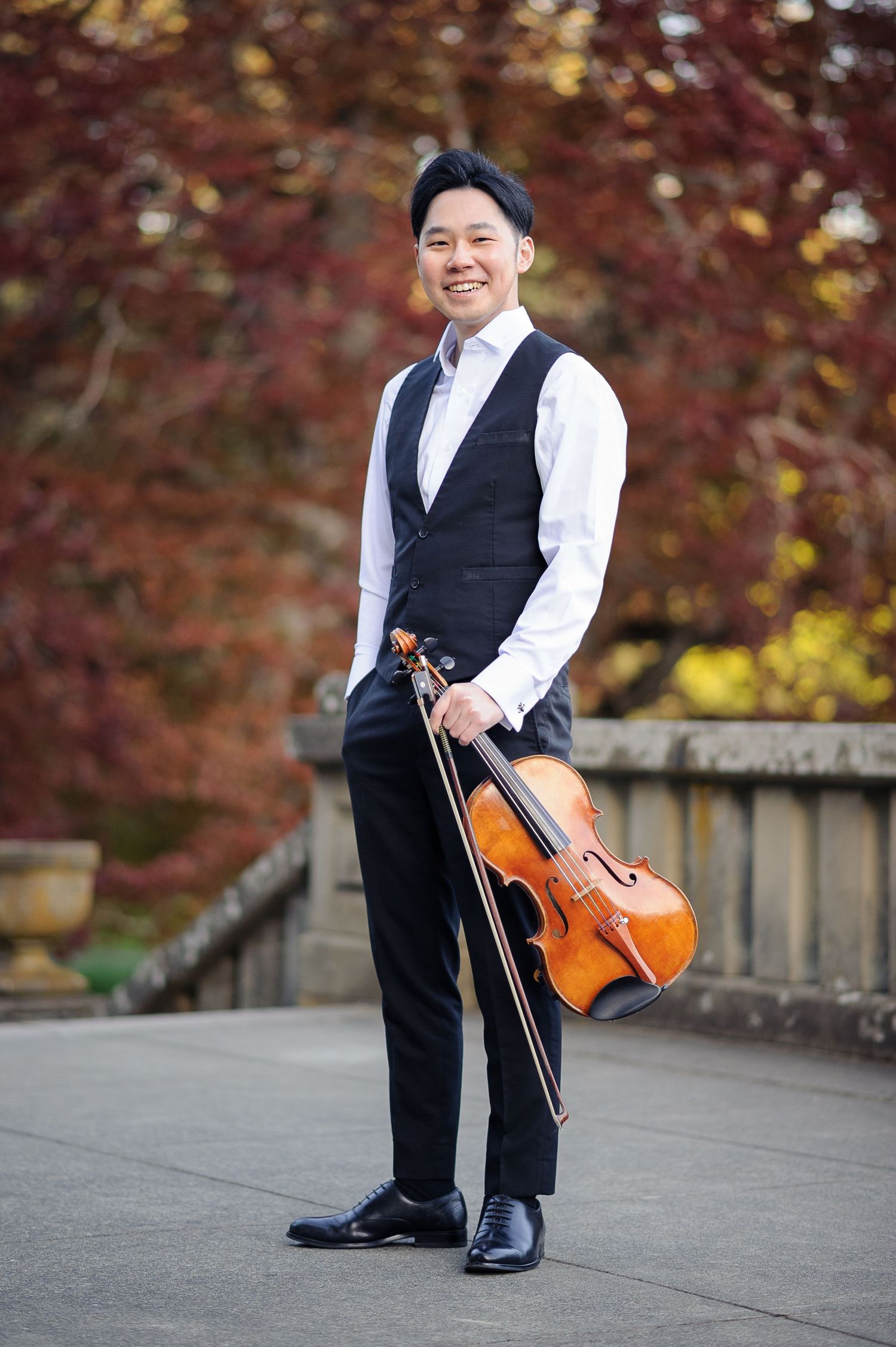 A man in an elegant vest smiles and holds a viola on a landing by some red trees.