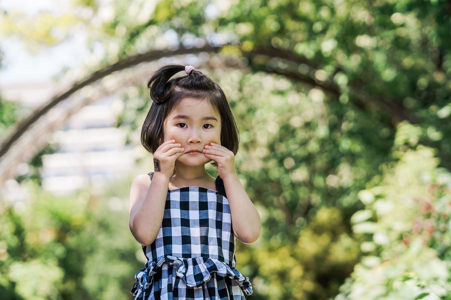 A young girl in a checkered sundress stands in a leafy garden and makes a funny face.