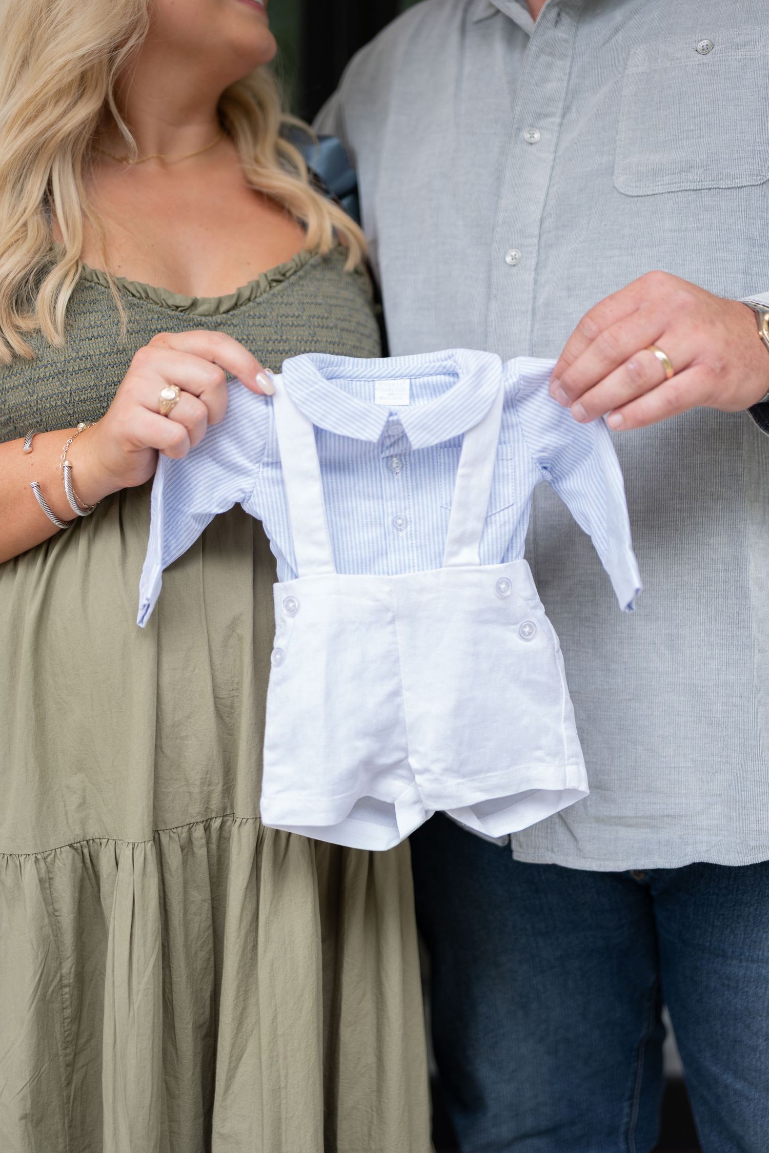 Couple holding up tiny white baby clothes outdoors.