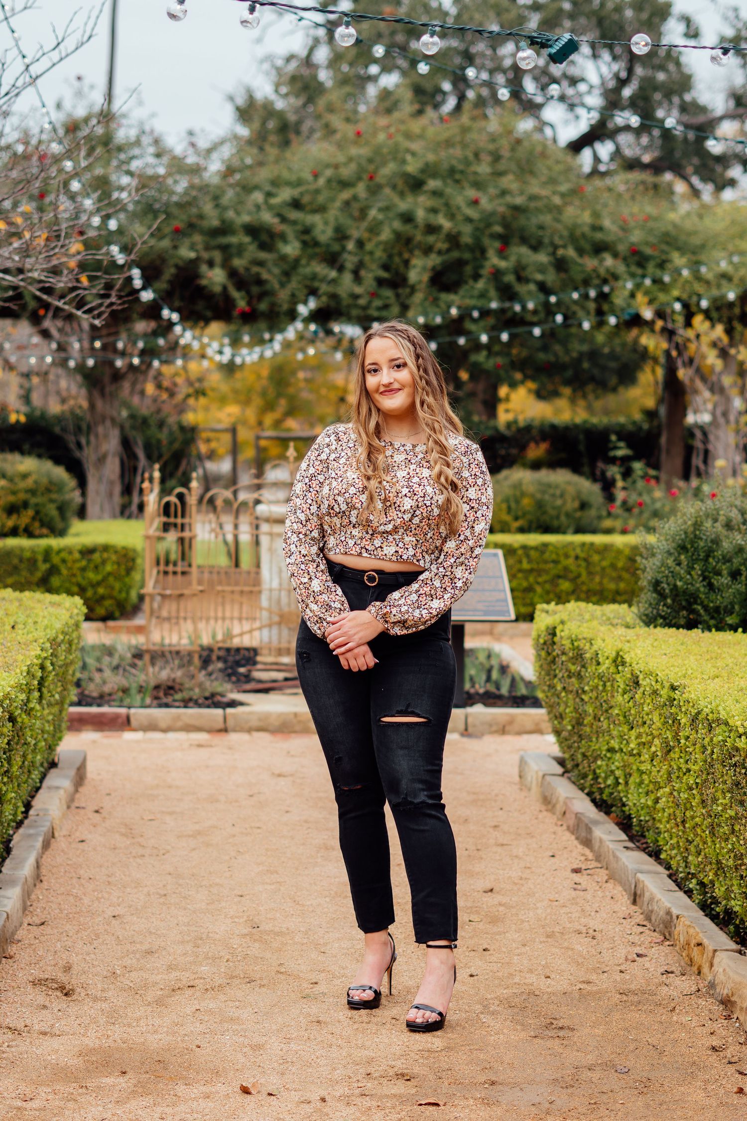 Person walking along a garden path lined with manicured hedges wearing a leopard print top and distressed jeans.