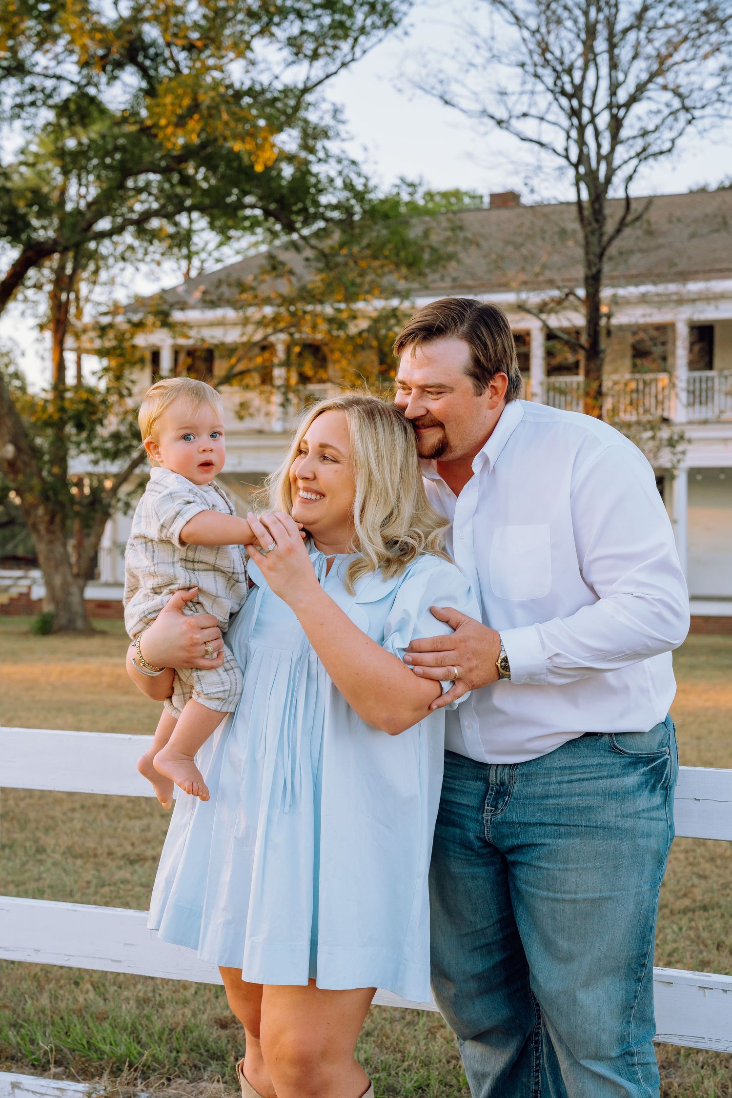 Family laughs together during a casual outdoor photo session by a white fence.