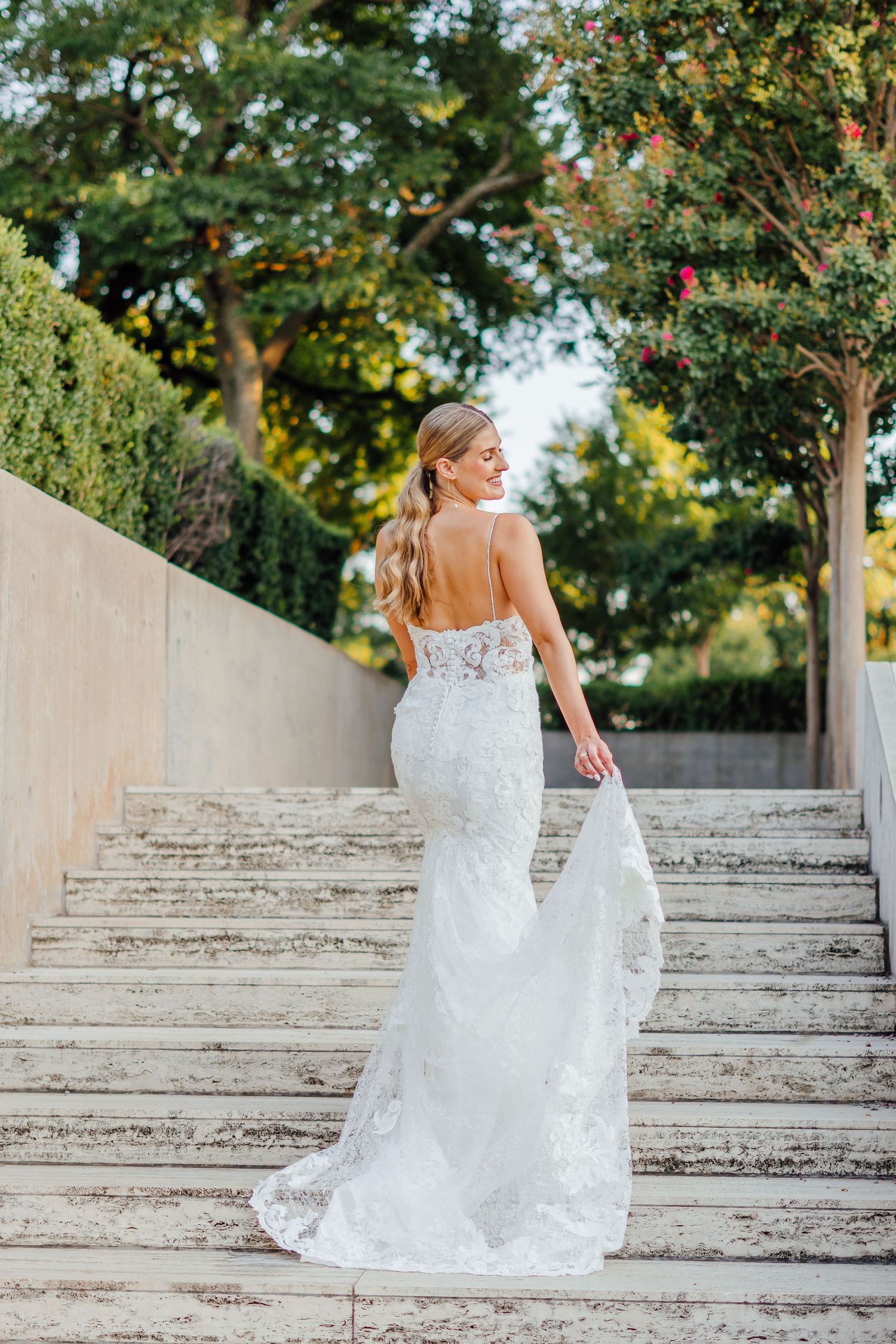 Elegant bridal portrait on stone steps with flowing white lace gown and garden backdrop.