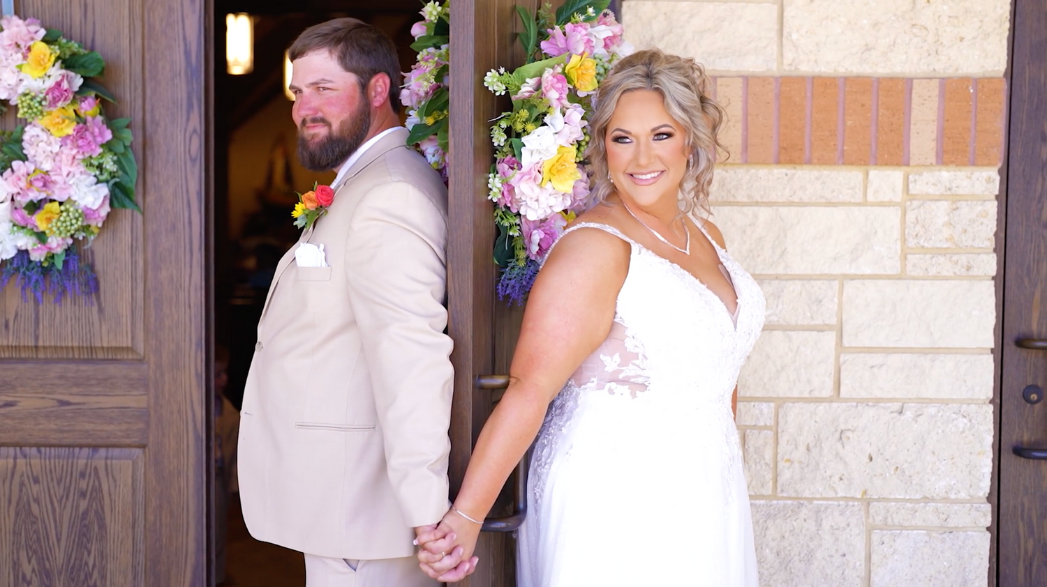 Wedding party poses near stone building with colorful floral arrangements and decorative arch.