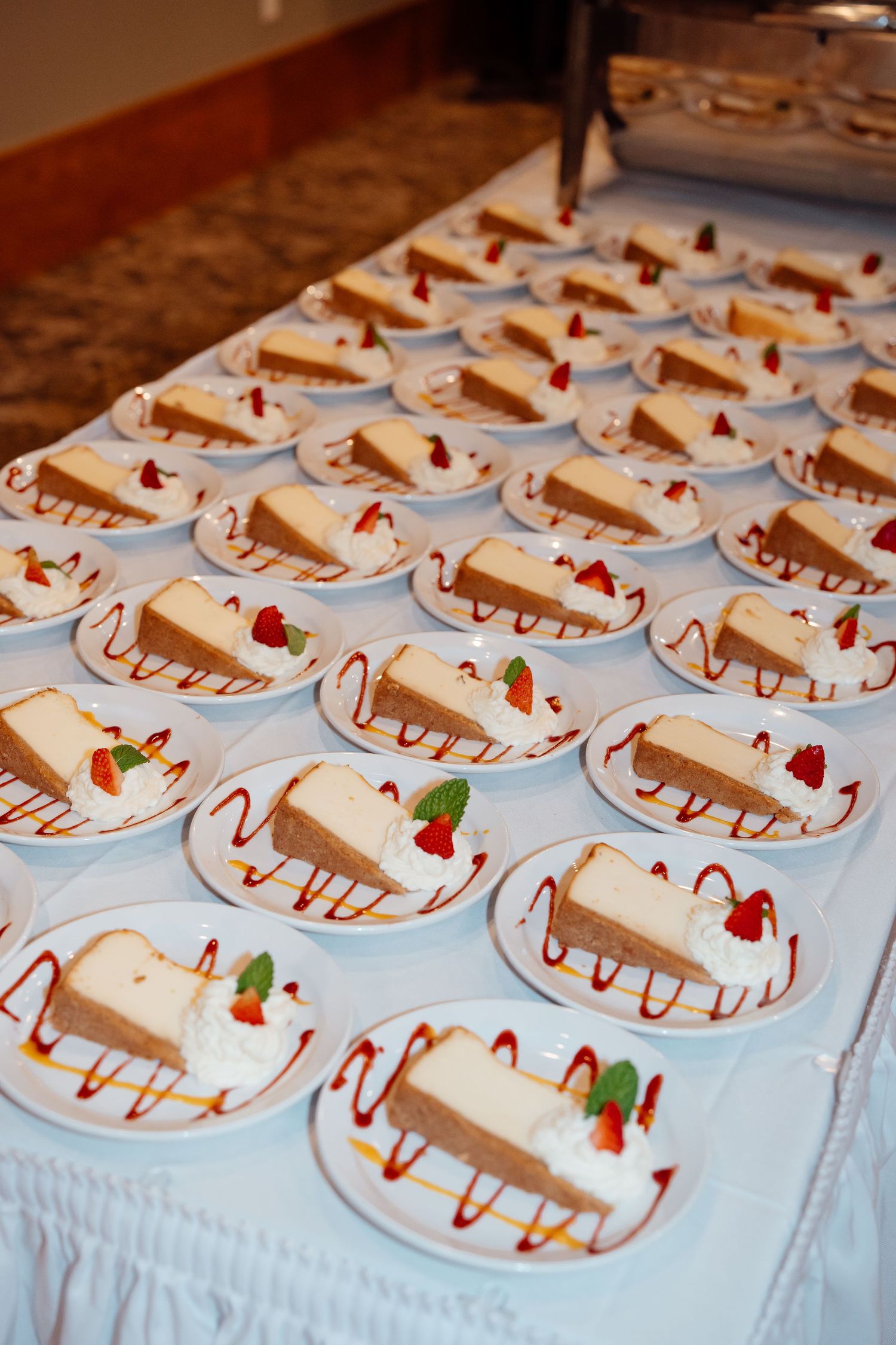 Rows of elegantly plated cheesecake desserts with raspberry sauce and mint garnish on white tablecloths.