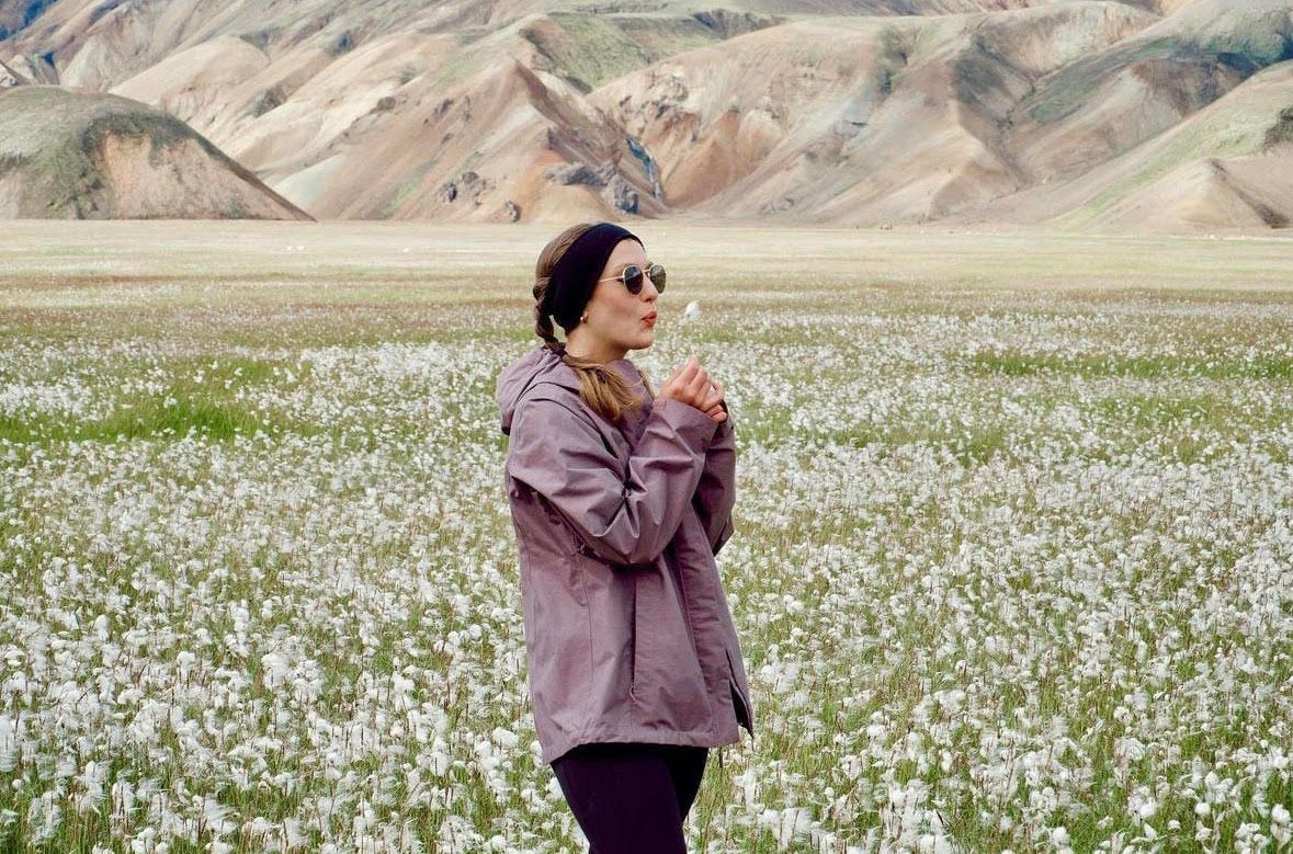 Person in purple jacket walking through field of white wildflowers with mountains in background.