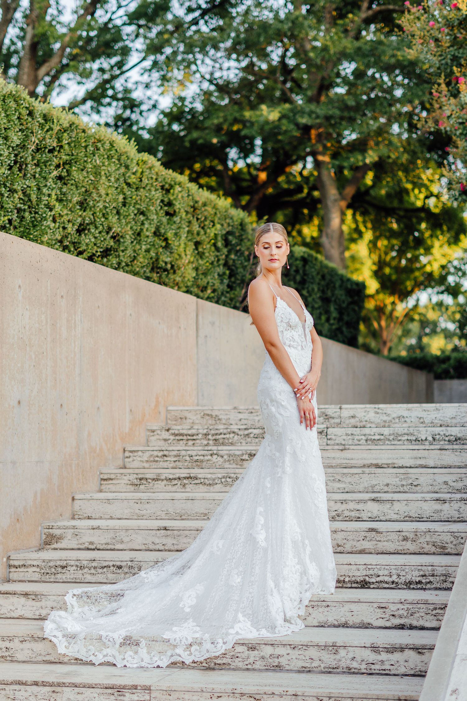A bride in a lace wedding gown poses elegantly on concrete steps outdoors.