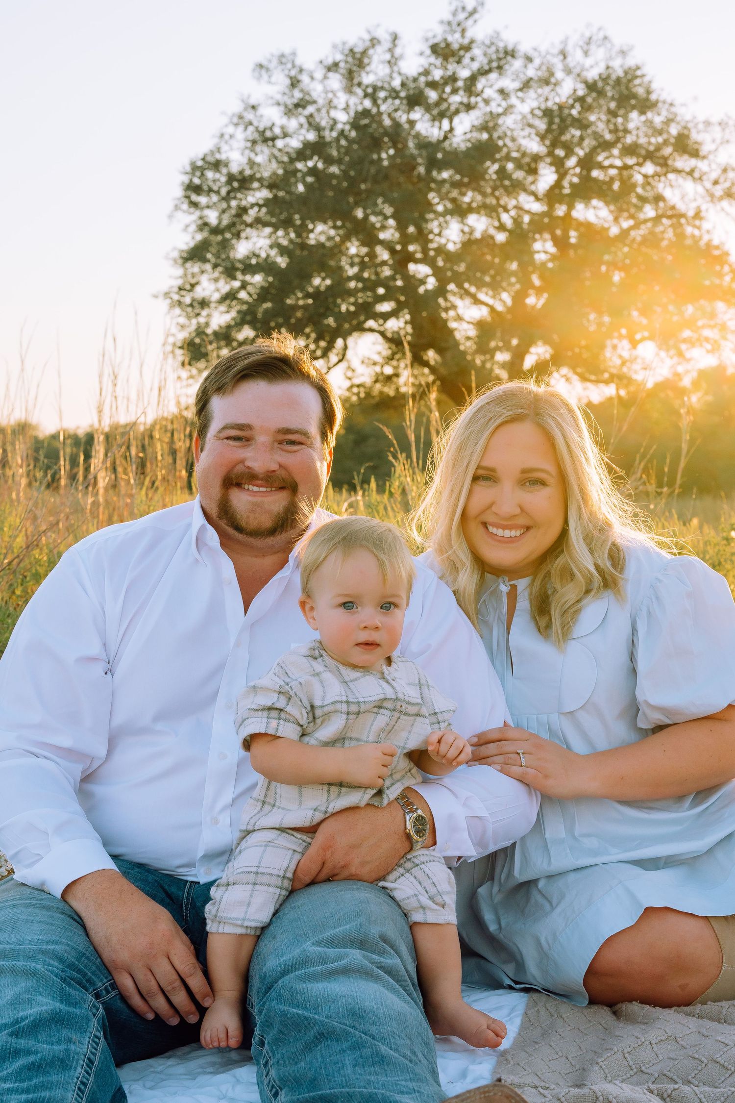 A family sits together on a blanket during golden hour with a tree silhouetted in the background.