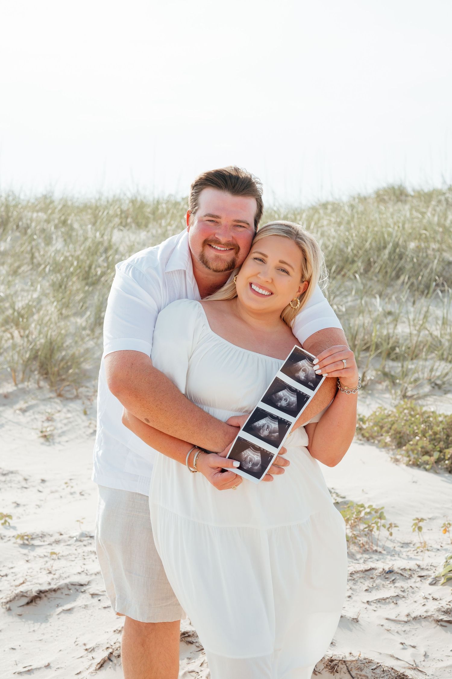 Couple on beach holding ultrasound photo while embracing in casual white outfits during pregnancy announcement photoshoot.