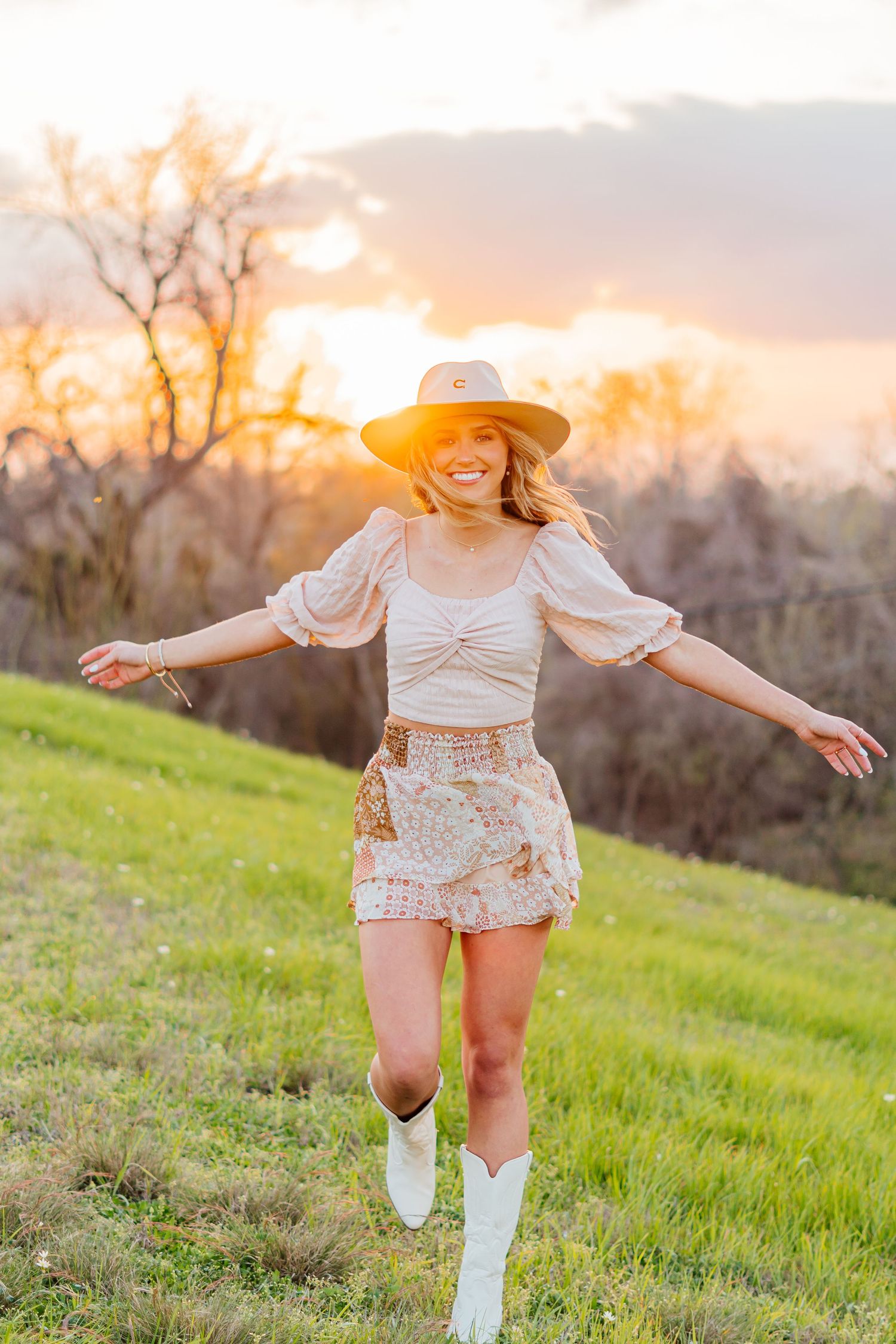 Person in white outfit and cowboy boots dances in grassy field during sunset.