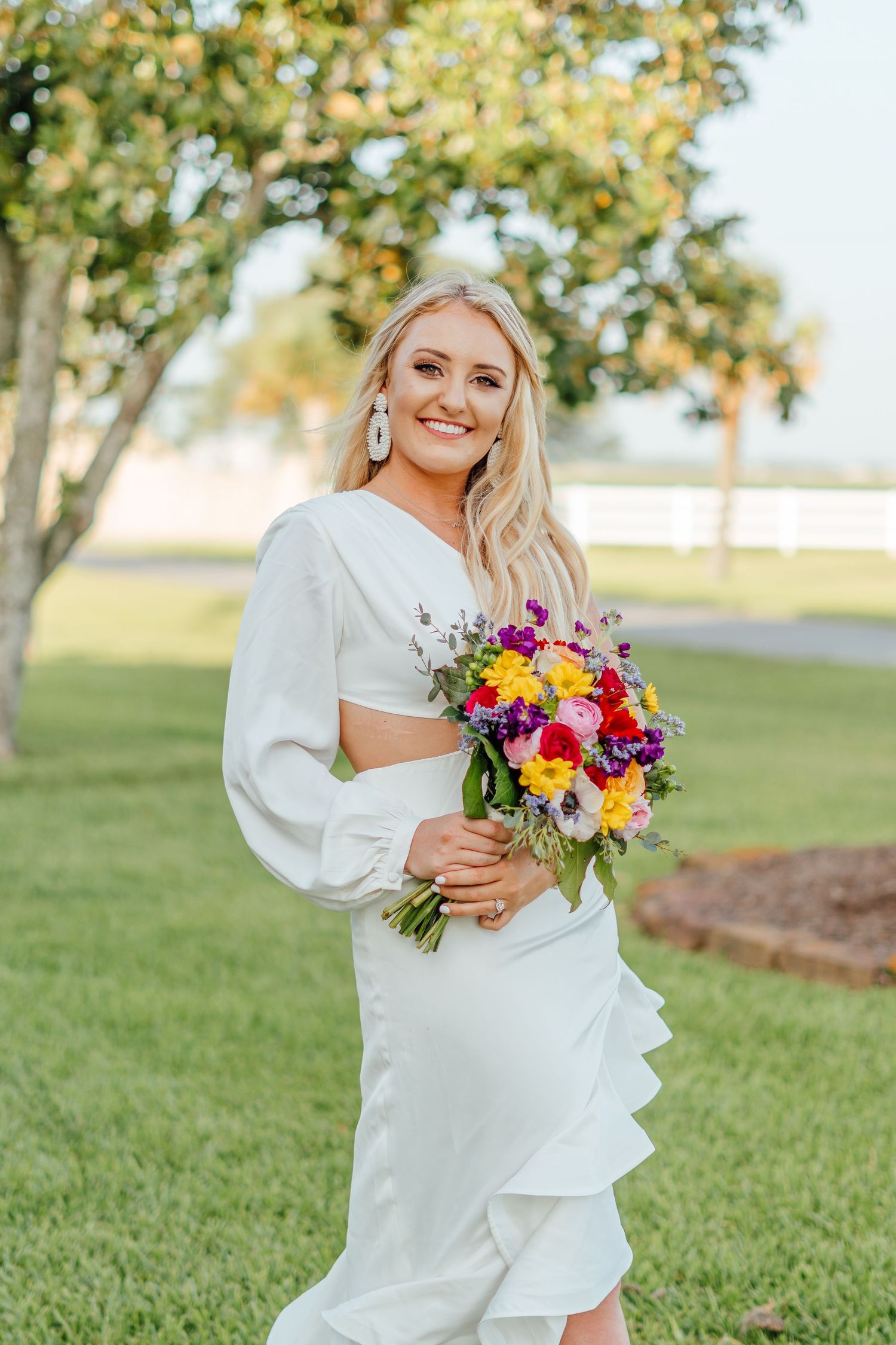 Bride in white dress holding colorful bouquet against green lawn and trees.