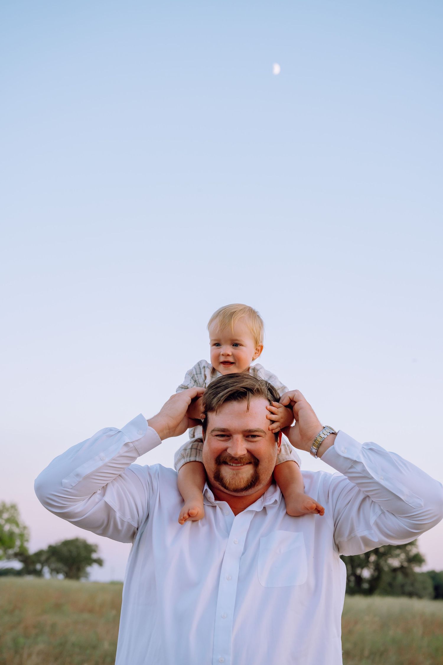 A toddler rides on shoulders outdoors at sunset with the moon visible in the sky.
