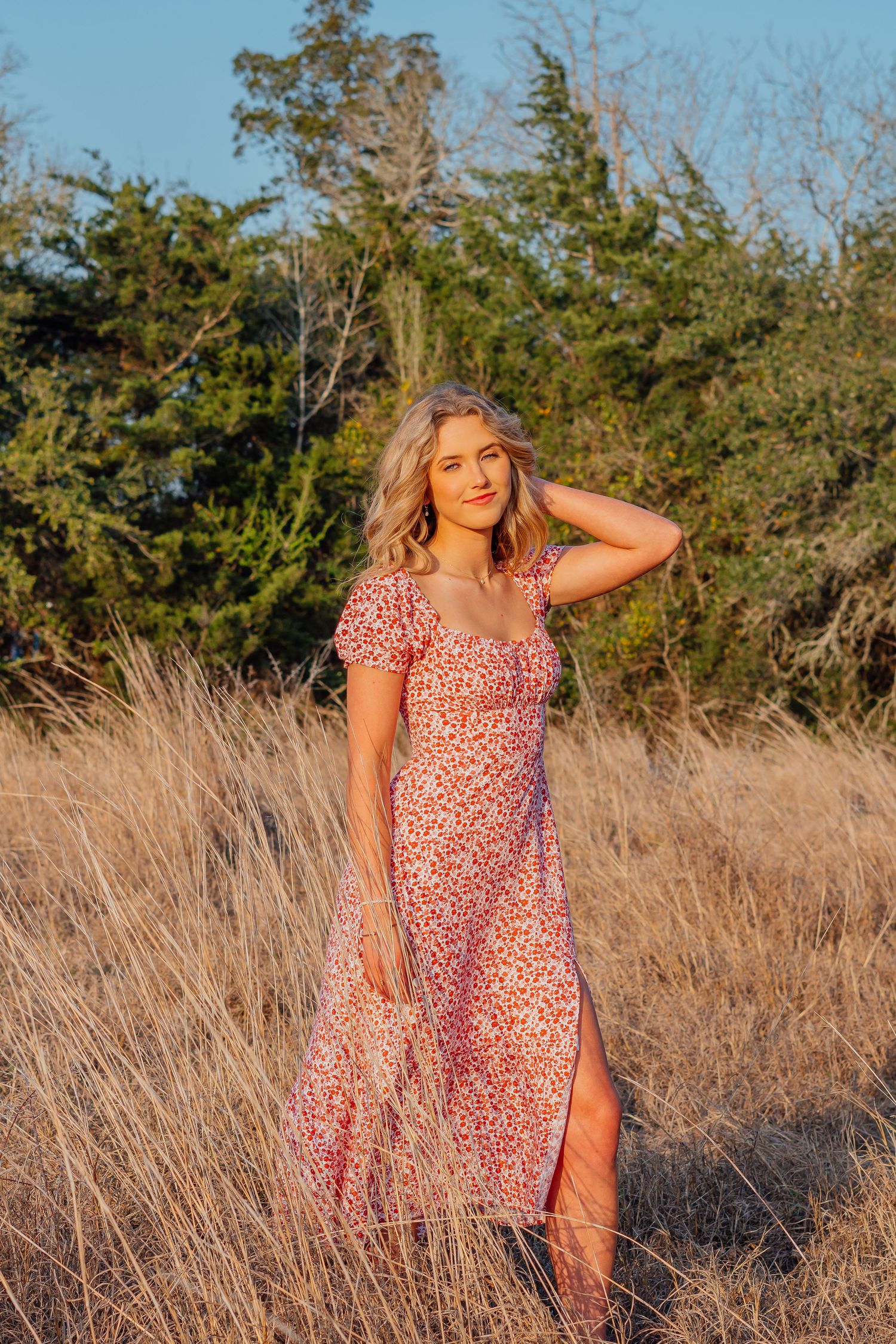 Person in floral dress poses in golden field during sunset with trees in background.