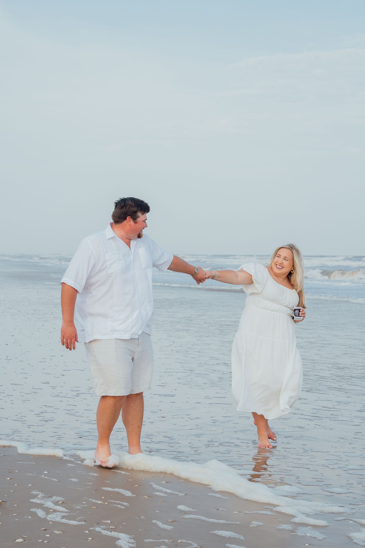 Couple playing in ocean waves during beach maternity photoshoot in white outfits.