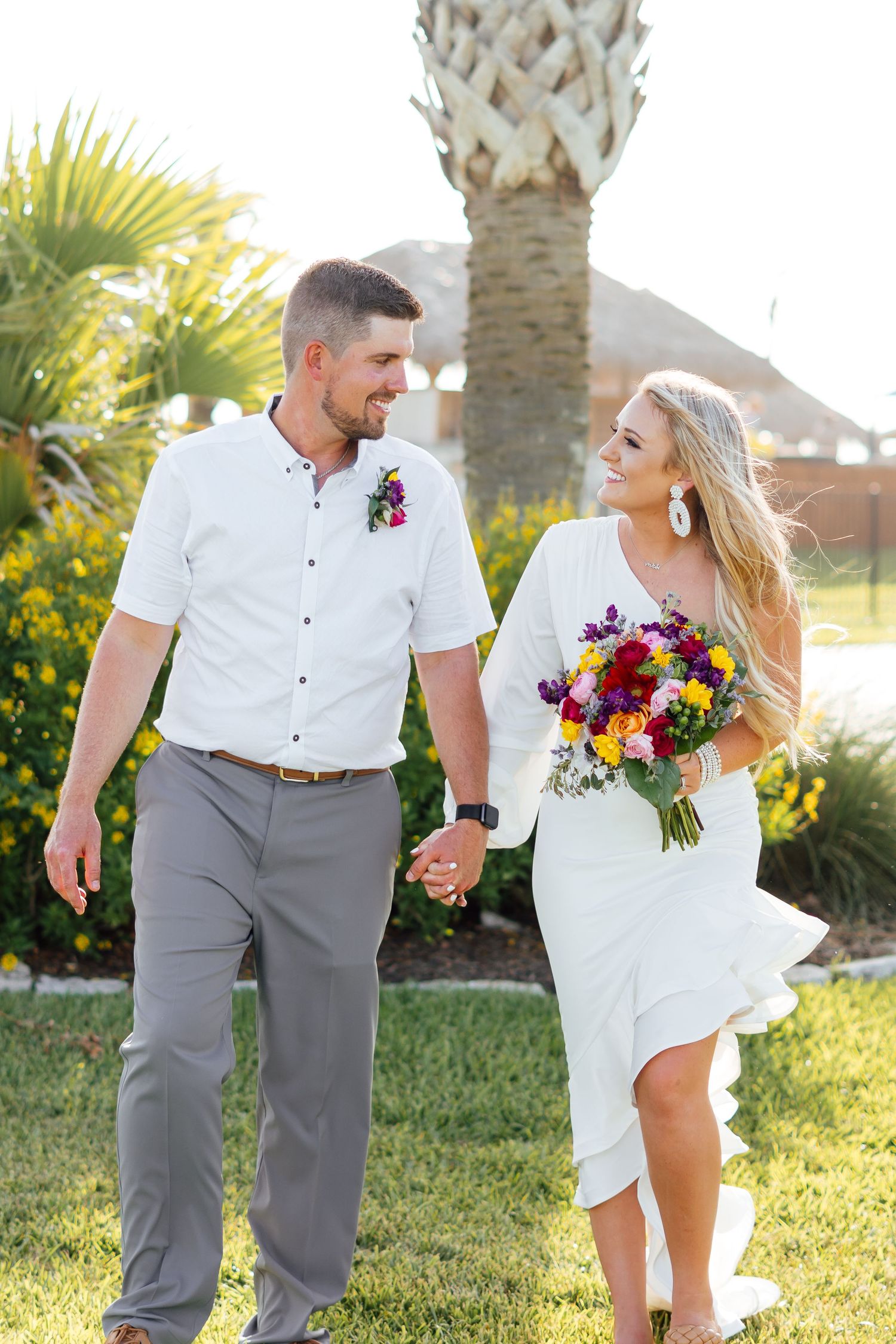Beach wedding photoshoot with couple in white and gray attire holding colorful flowers.
