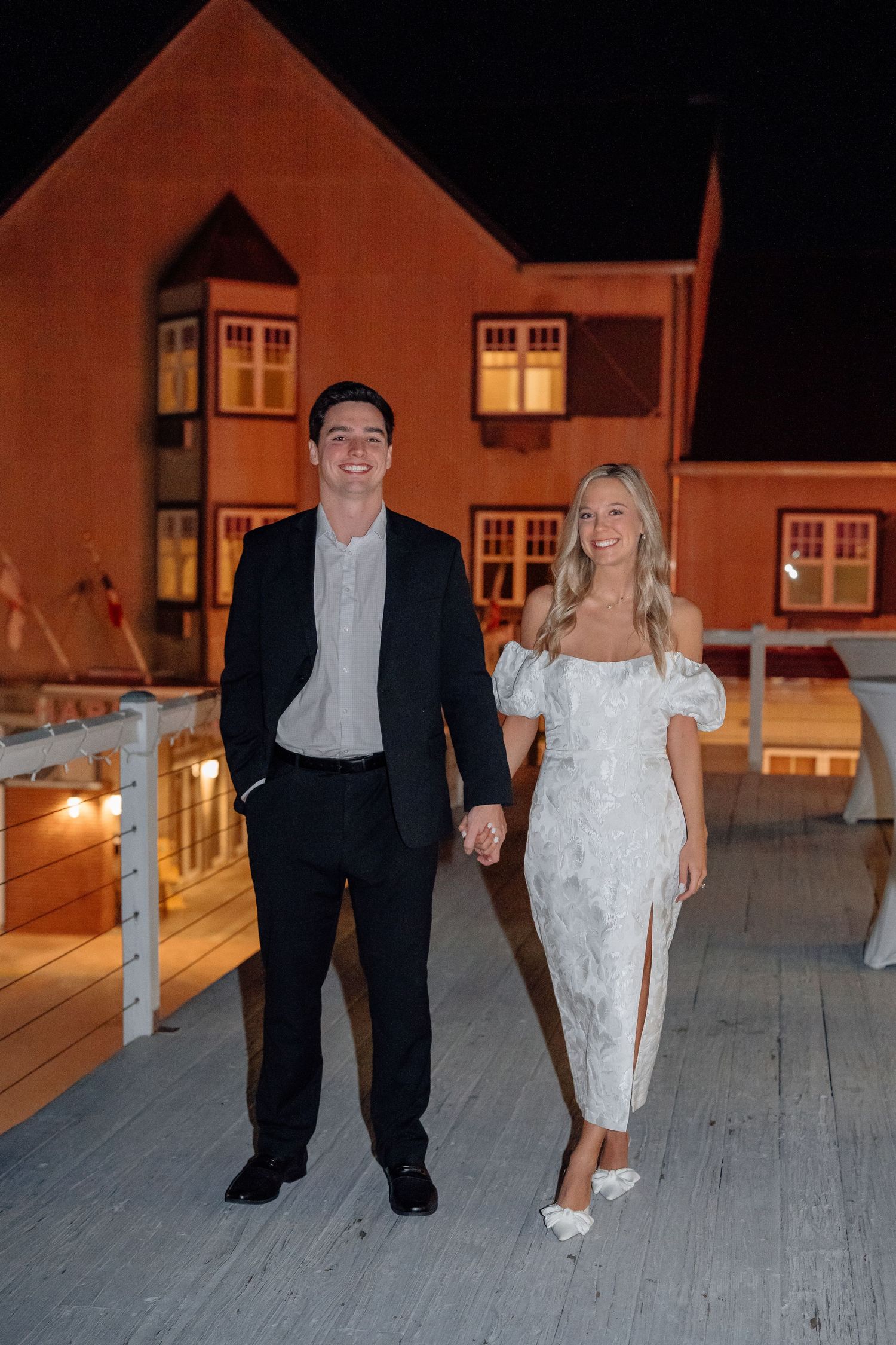 Couple walking together on a wooden deck at night with illuminated building in background.