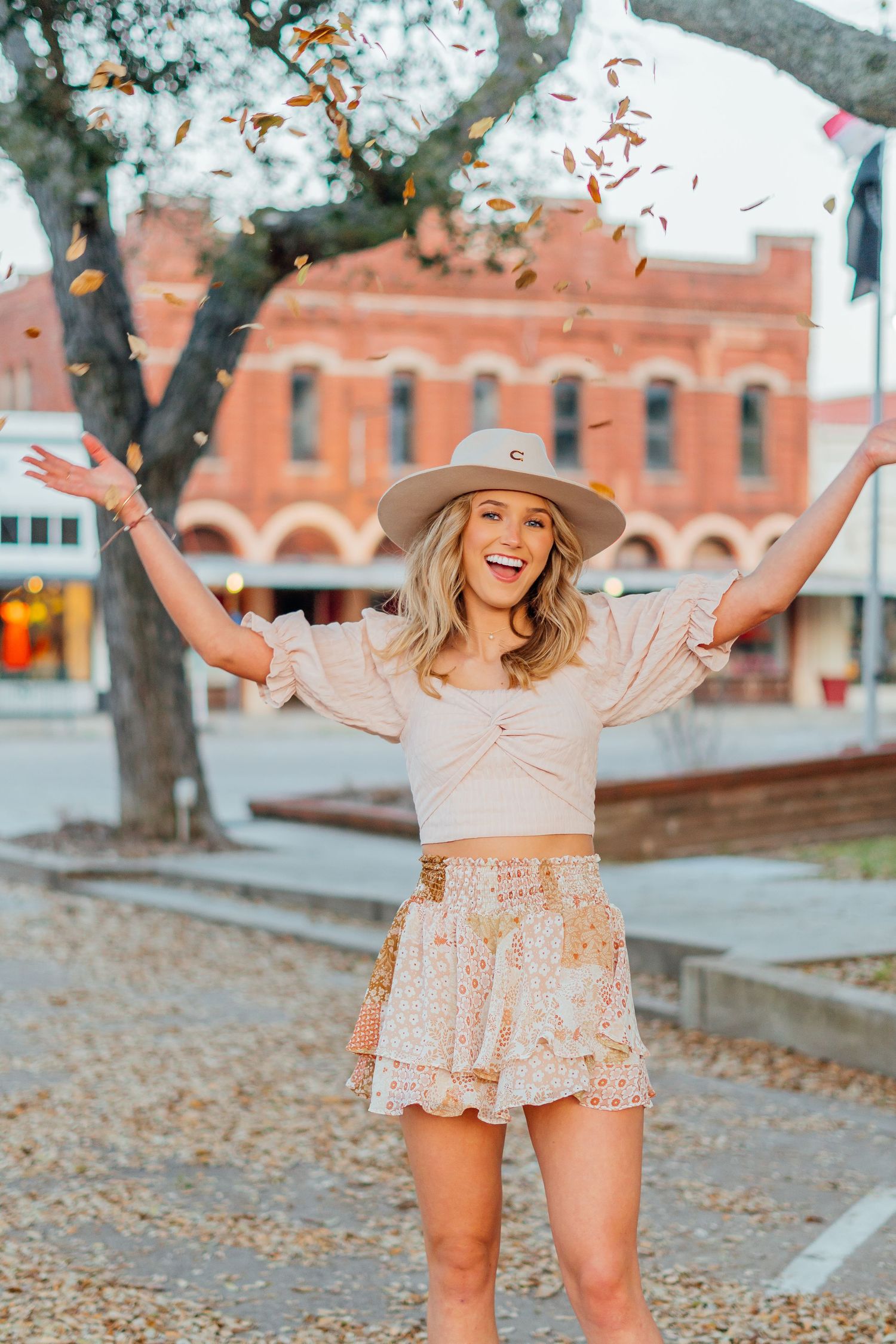 Woman in beige dress and hat joyfully throws autumn leaves in the air near brick buildings.