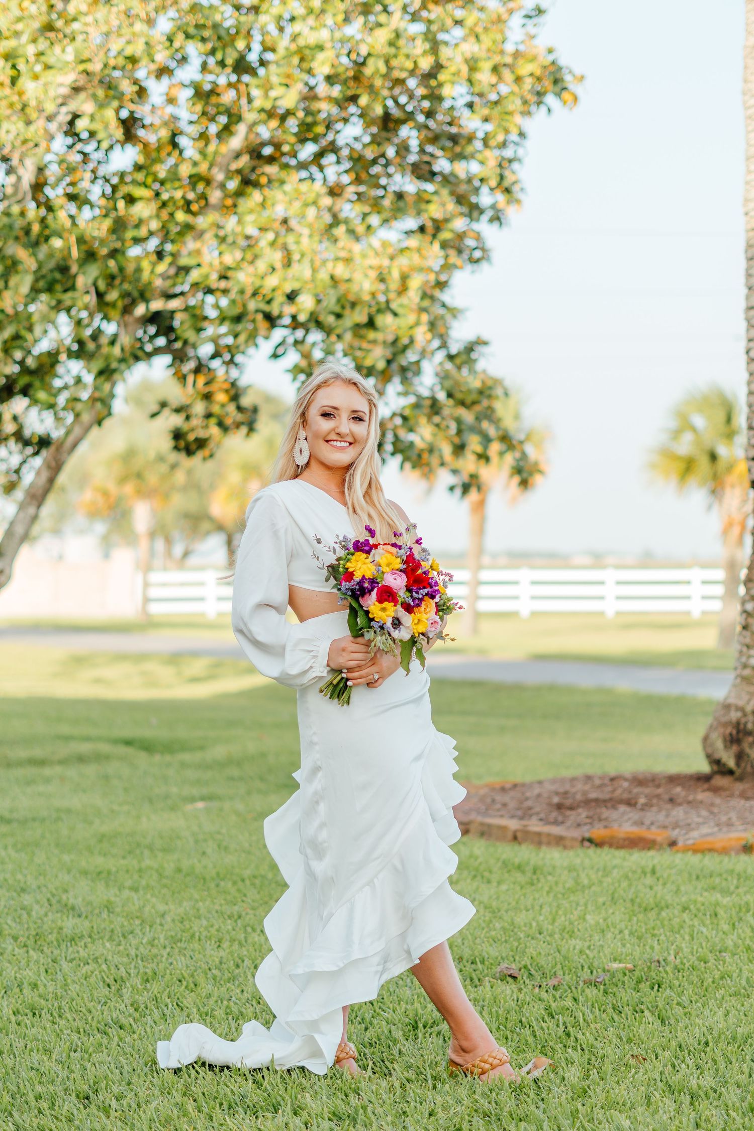 Bride walking on grass holding vibrant bouquet with white fence and trees in background.