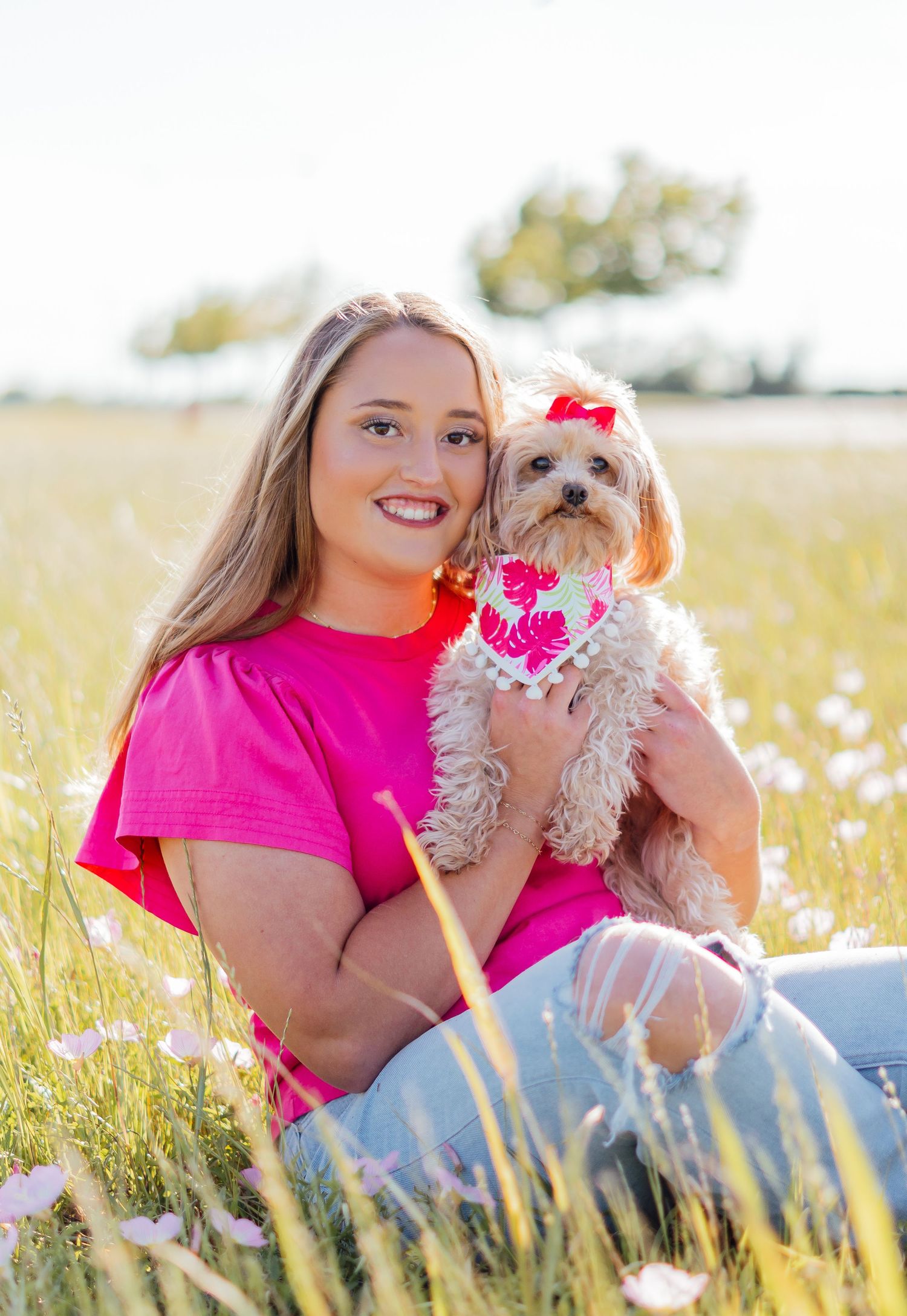 Person in bright pink shirt holding small dog in sunny wildflower field.