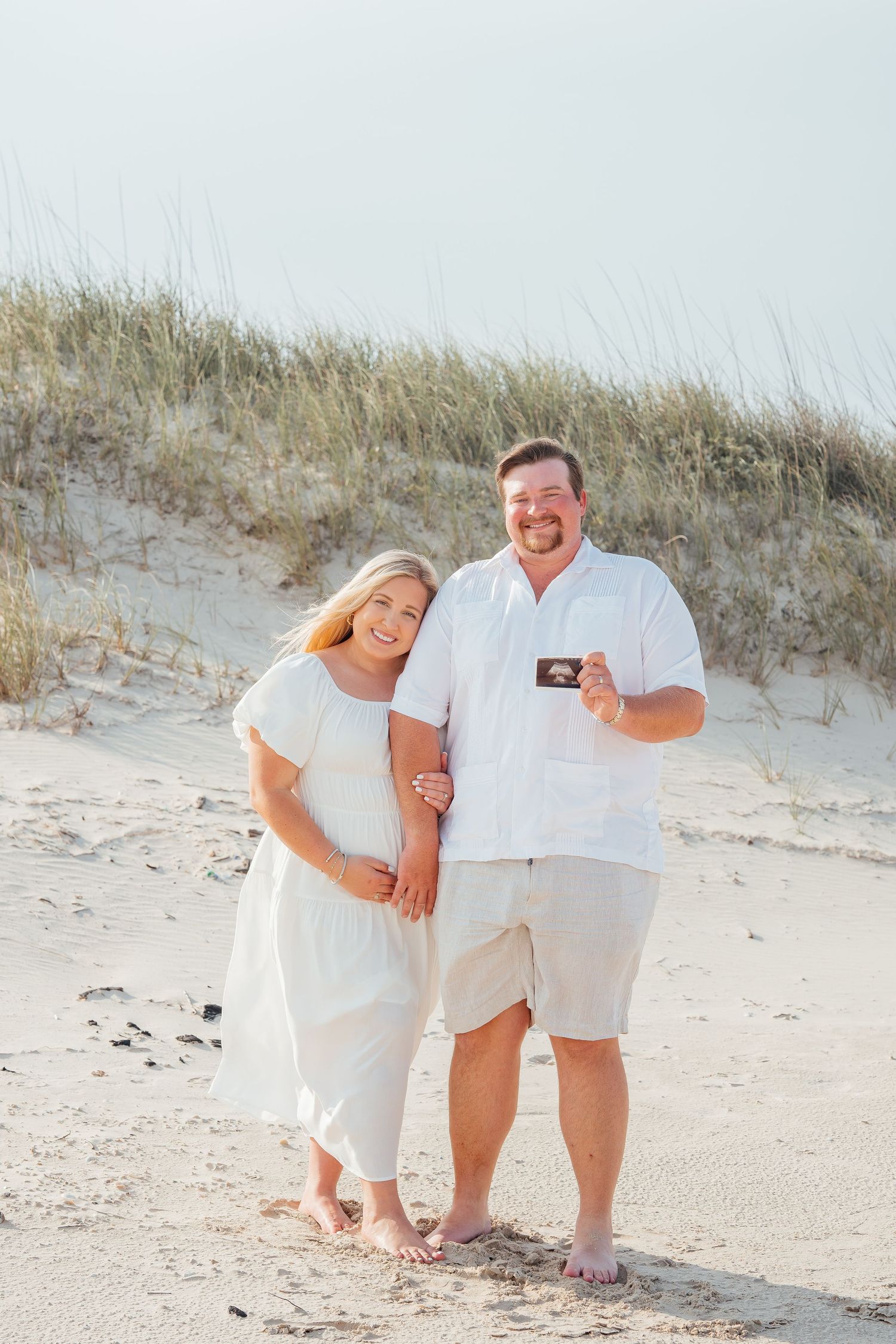 Happy couple in white outfits sharing ultrasound photo on beach dunes.