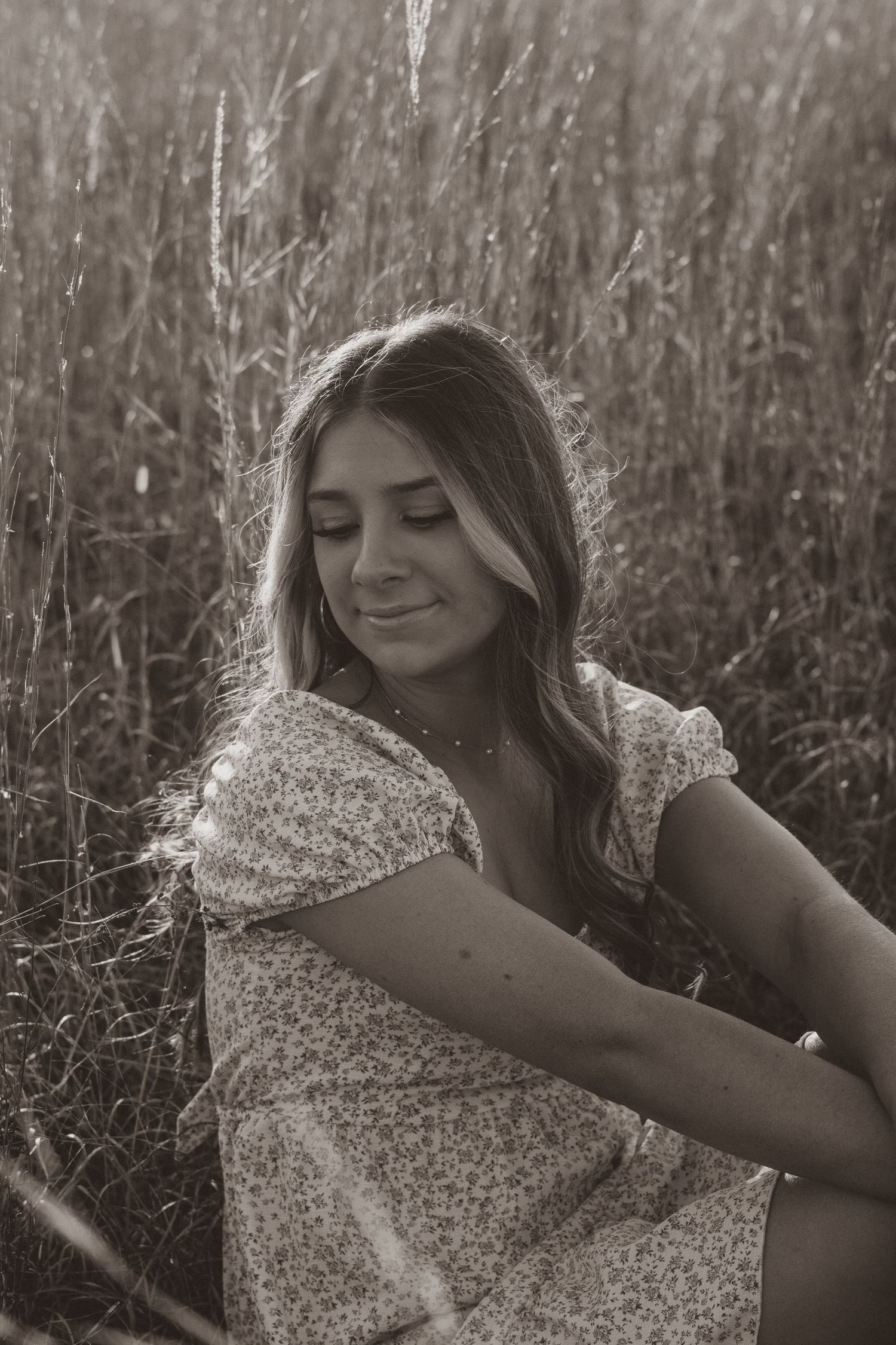 Woman in floral dress smiling with eyes closed in a sunlit field of tall grass.