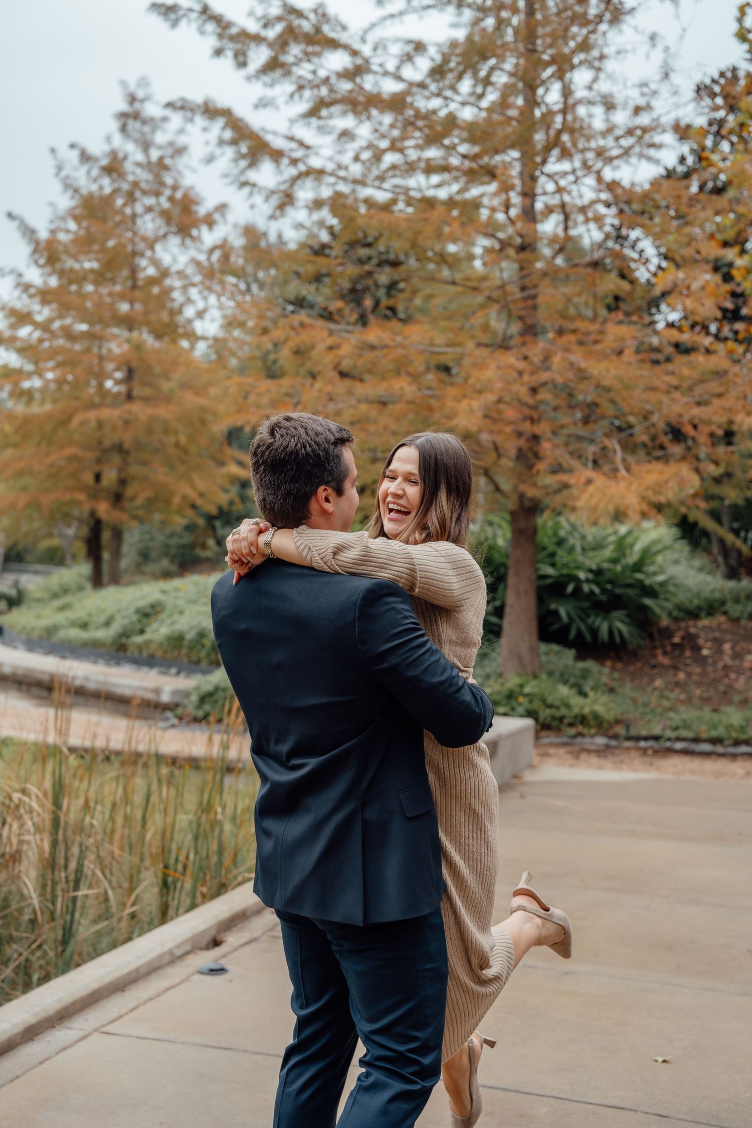 A playful couple spins and dances together on a pathway surrounded by fall foliage.