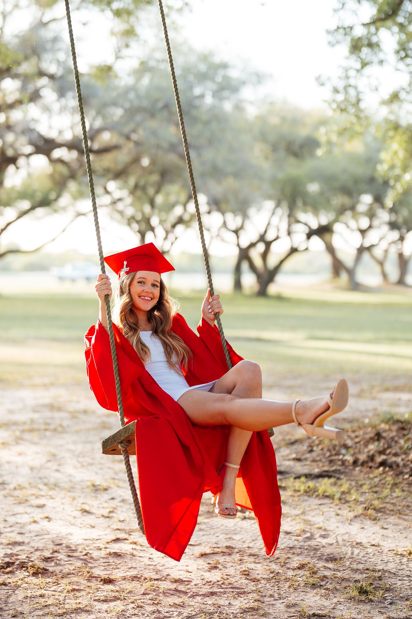 Graduate in red cap and gown swings joyfully under oak trees during sunset photo session.