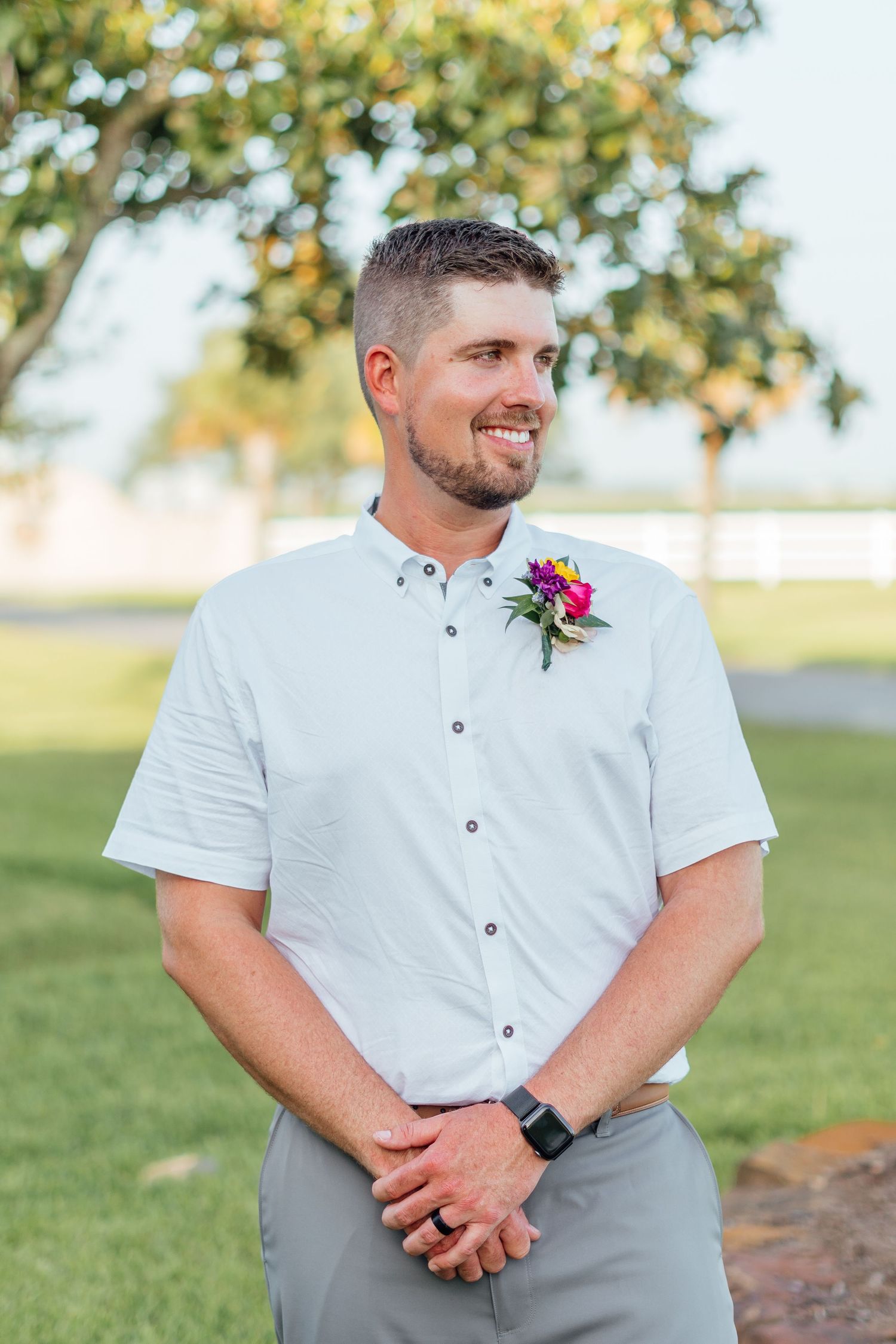 Man in white dress shirt wearing a pink boutonniere smiles outdoors during a wedding ceremony.
