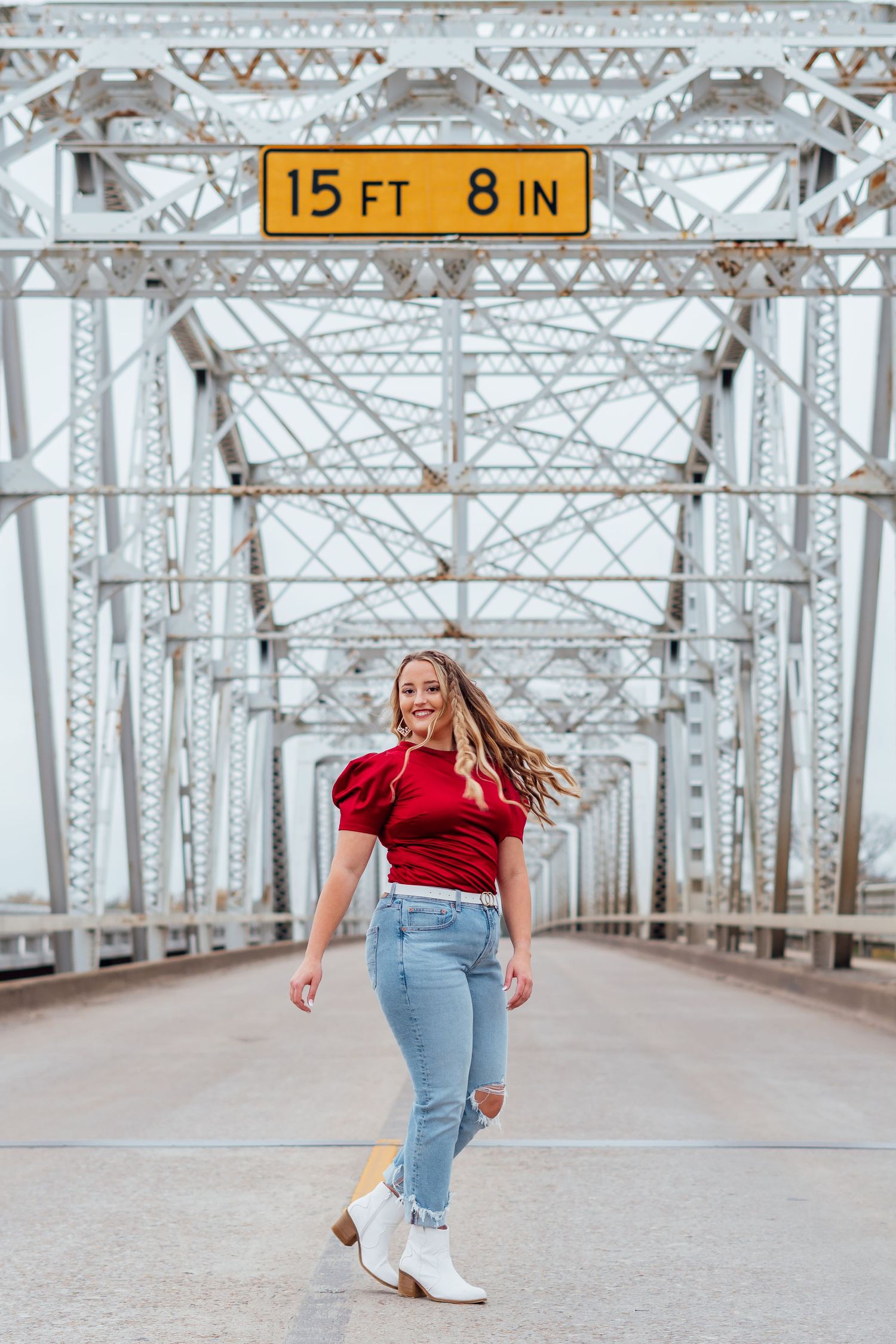 Playful poses on metal bridge walkway wearing red top and jeans with white boots.