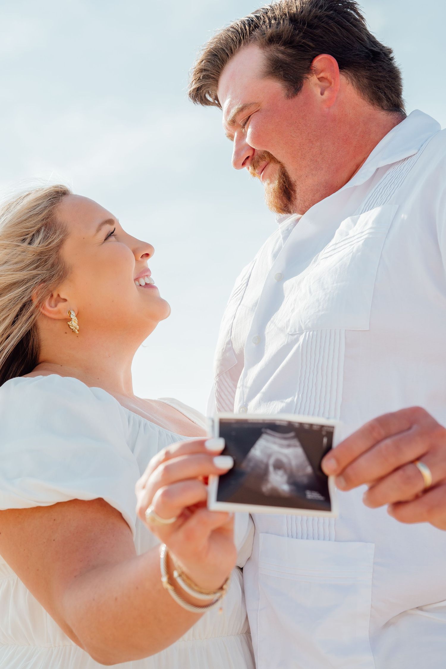Couple in white sharing joyful moment with ultrasound photo at beach.