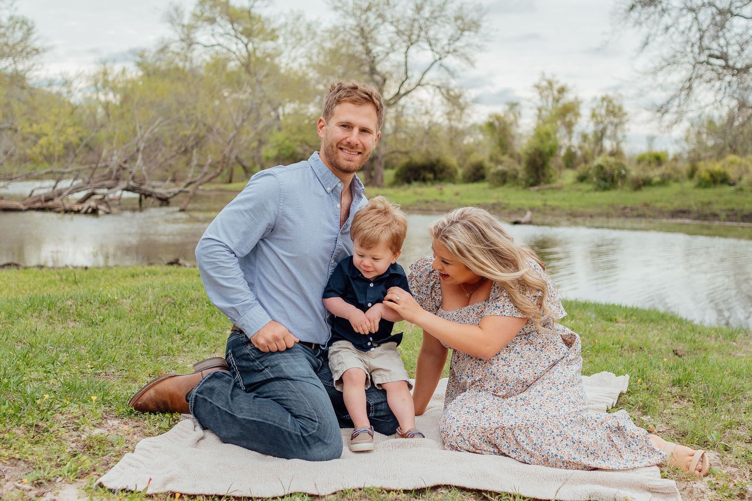 Family lying on blanket during outdoor photoshoot near water with natural landscape in background.