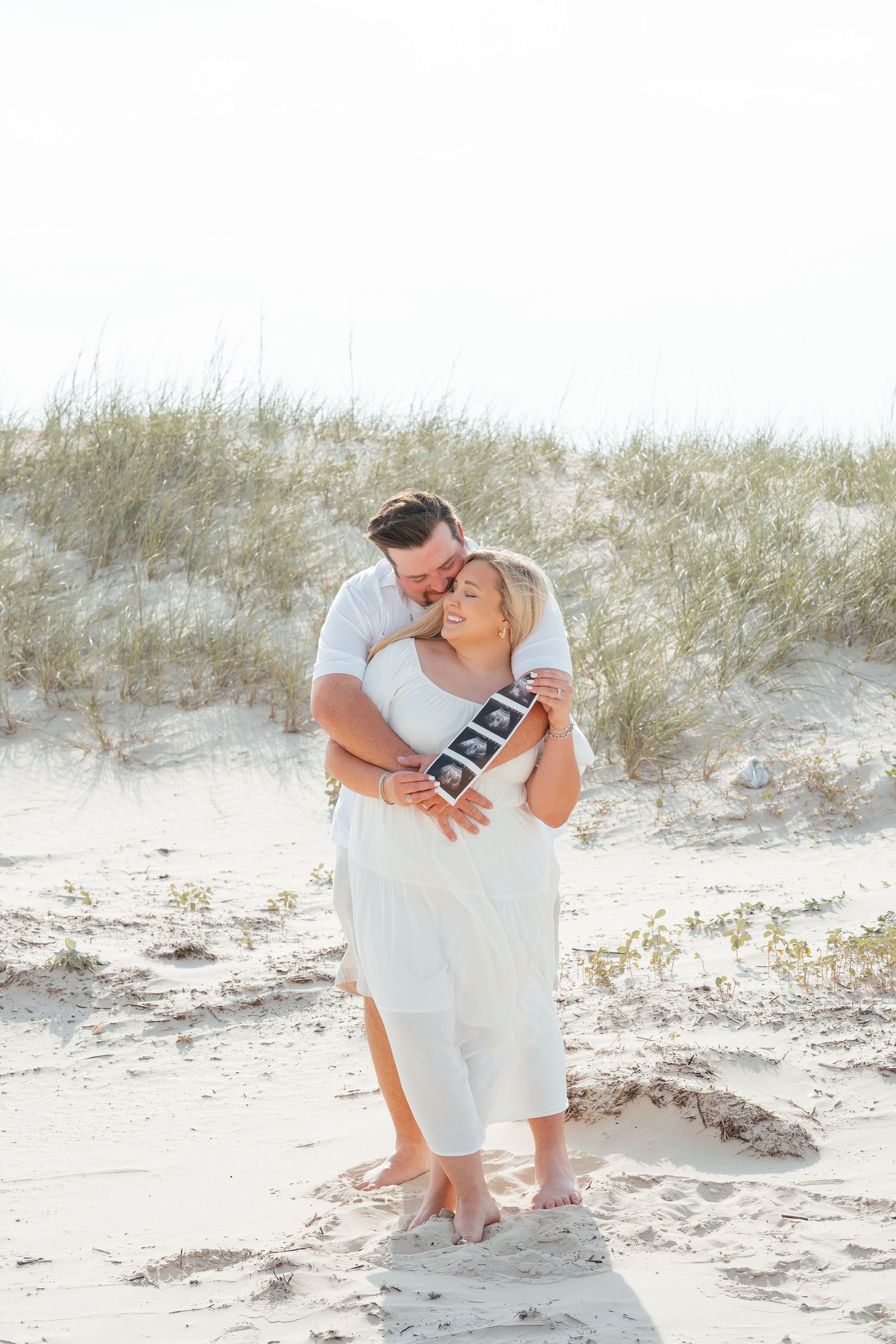 Happy couple playfully posing on sandy beach during pregnancy announcement session.