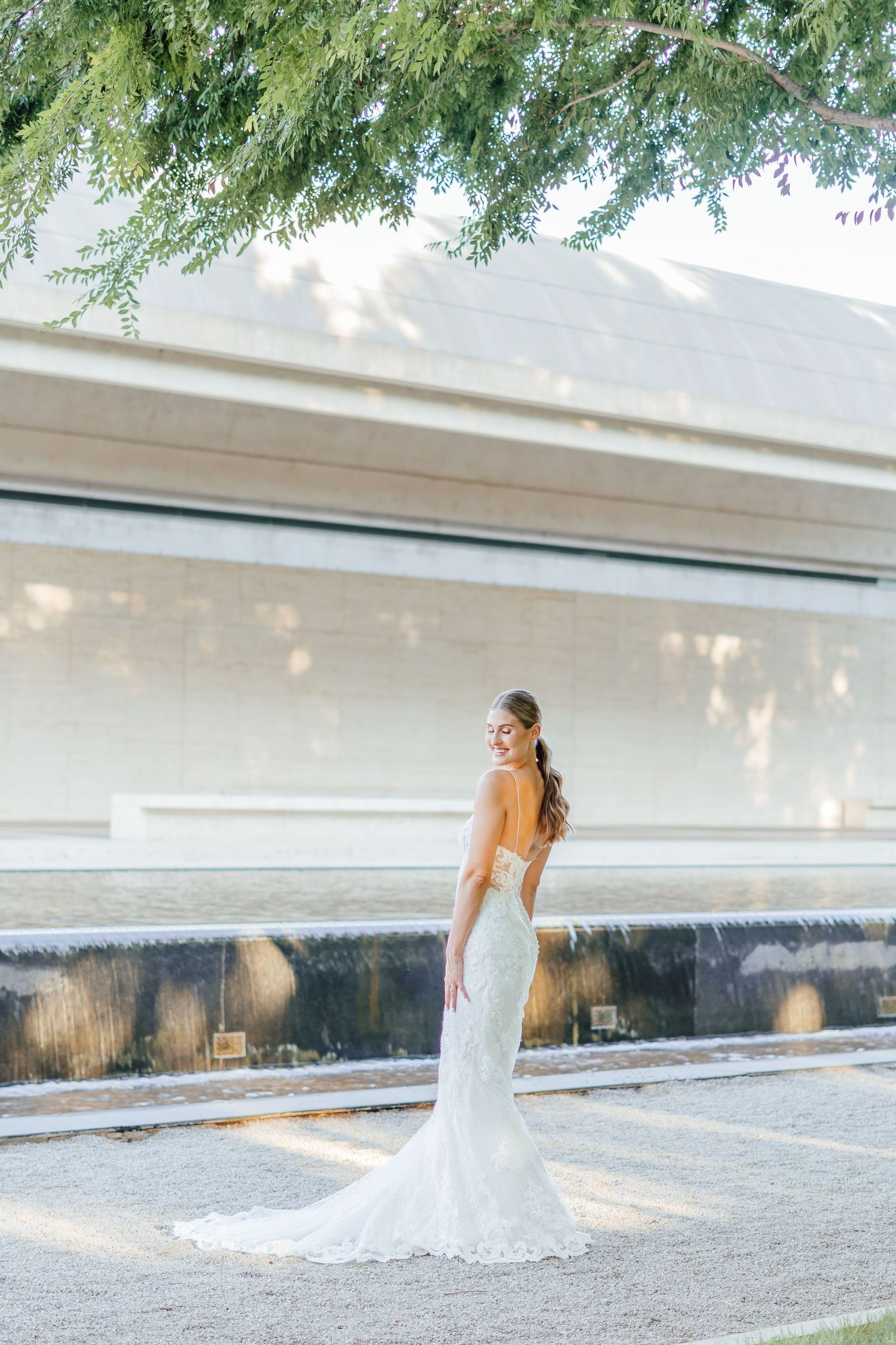 A bride in a flowing white dress poses gracefully by a modern water feature.