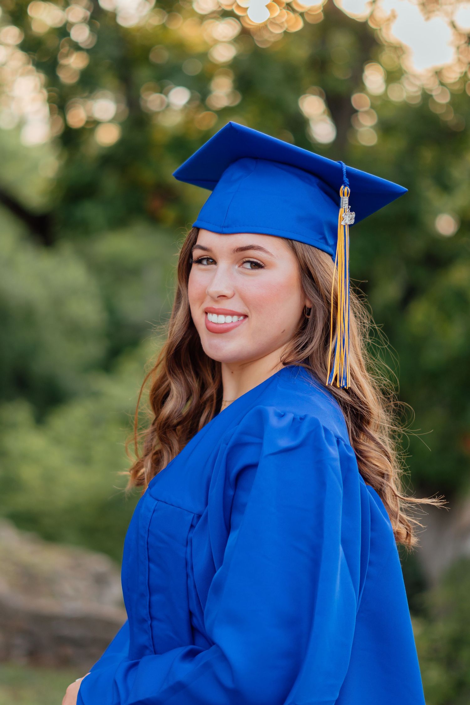 Graduate in royal blue cap and gown poses outdoors with natural bokeh background.