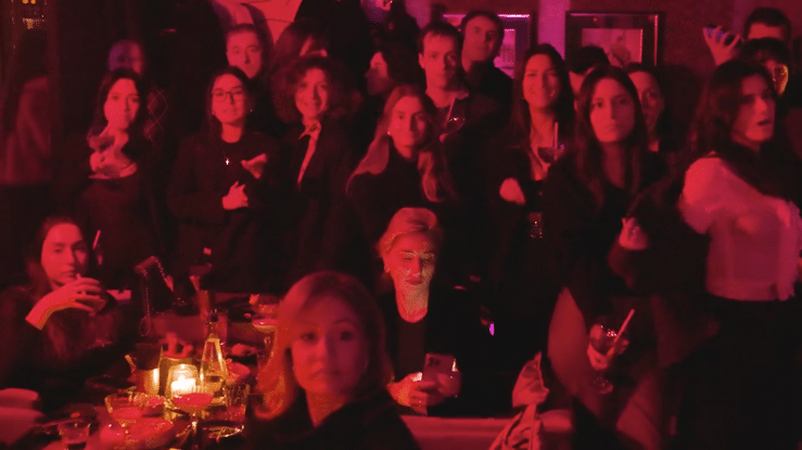 Concert audience illuminated by red stage lights watches a performance in a dimly lit venue.