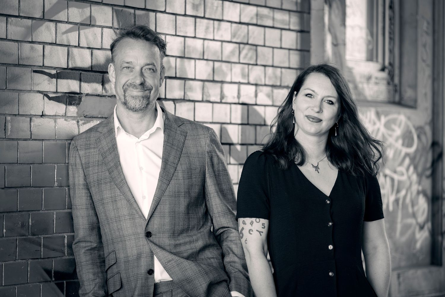 Black and white photo of a man in a blazer and a woman with tattoos smiling in front of a brick wall.