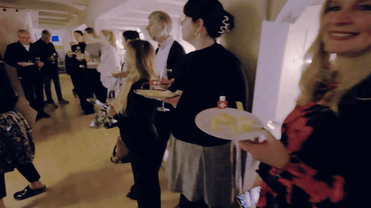 A group of people gather in a crowded indoor space with light colored walls and floors for a social event.